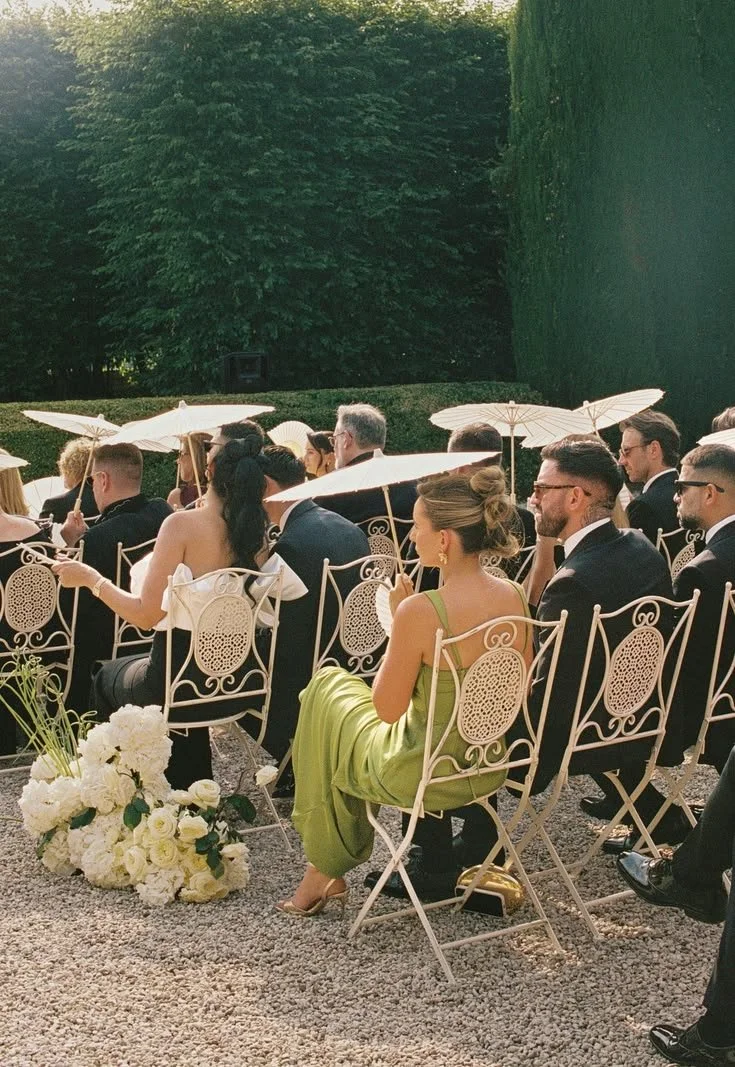 Guests seated outdoors at a wedding or special event, with some holding white parasols, surrounded by greenery, with flowers on the ground.