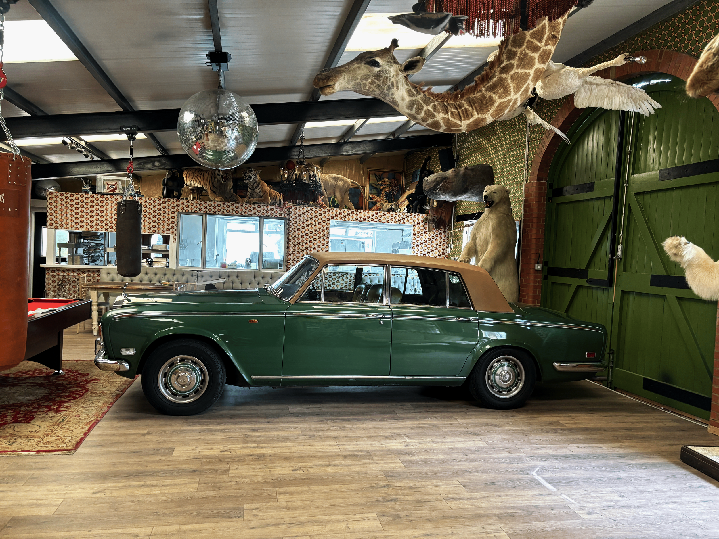A vintage green car with a tan roof parked indoors in a room decorated with animal pelts, statues, and mounted animal heads including a giraffe, bear, tiger, and several tigers.