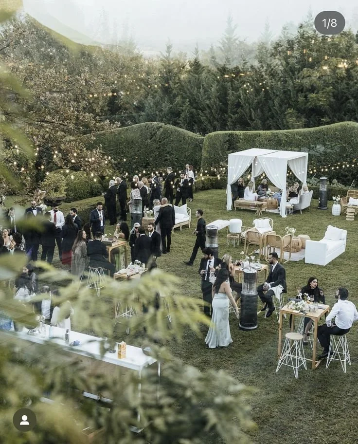 An outdoor gathering or party with people mingling and socializing on a lawn, decorated with string lights, tents, and various outdoor furniture, surrounded by tall hedge bushes and trees.