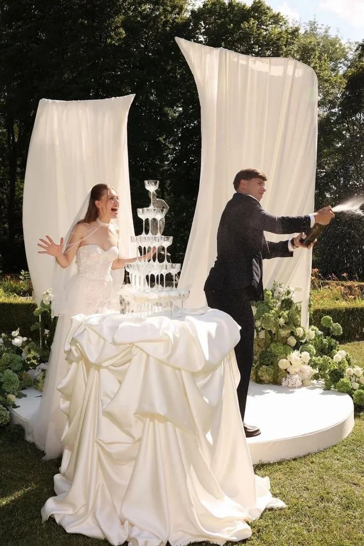 A bride and groom celebrating outdoors on their wedding day. The bride is wearing a white gown with a veil, and the groom is in a dark suit. They are using champagne to spray each other, with the bride laughing happily. There is a tiered champagne to