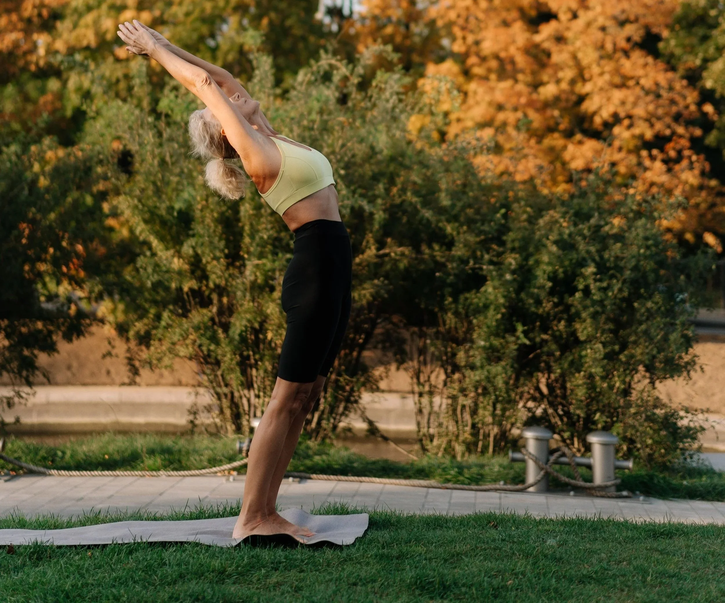 Older woman practicing yoga outdoors in a park during autumn, standing in downward dog pose on yoga mat.