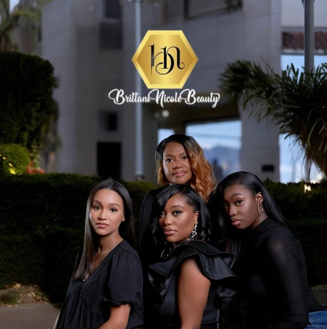 A logo with a gold hexagon and black stylized letter 'N' above the text 'Brittan Nicole Beauty,' and four women with dark hair dressed in black standing outdoors in front of greenery and buildings.