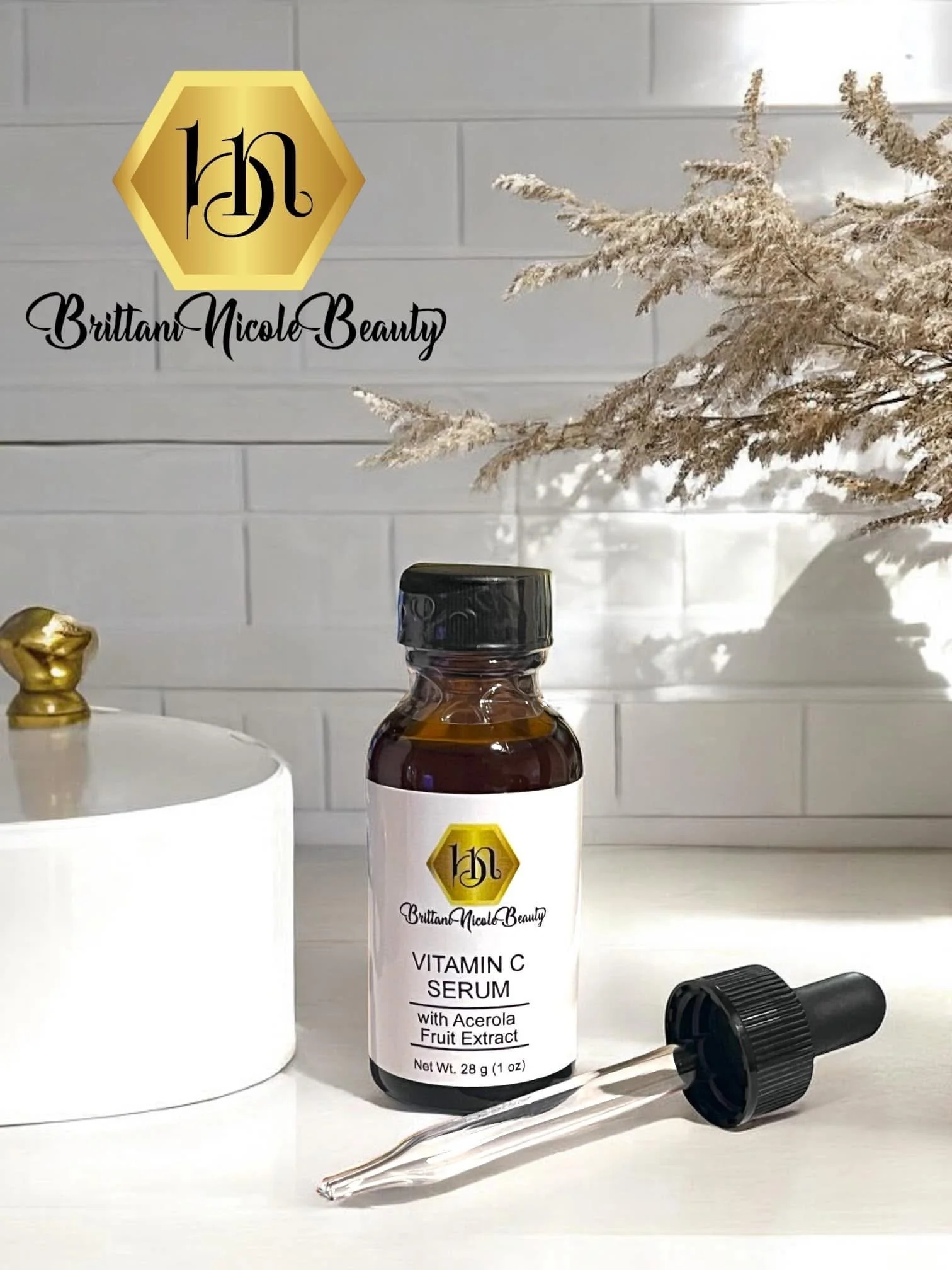A bottle of Brittany Nicole Beauty Vitamin C Serum with Acerola Fruit Extract placed on a white surface, alongside a black dropper cap, a white container, and a gold decorative object, with a background of a white tiled wall and dried decorative bran