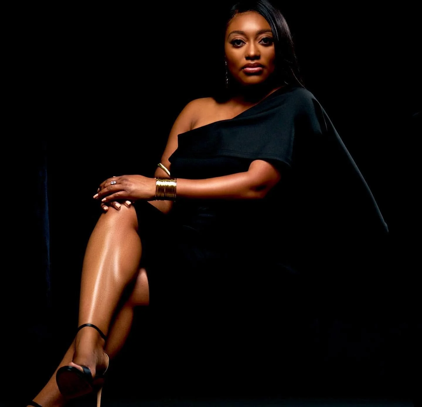A woman wearing a black off-shoulder dress, sitting with one leg crossed over the other against a dark background. She is accessorized with gold jewelry, including a bracelet and rings, and has long, dark hair and makeup.