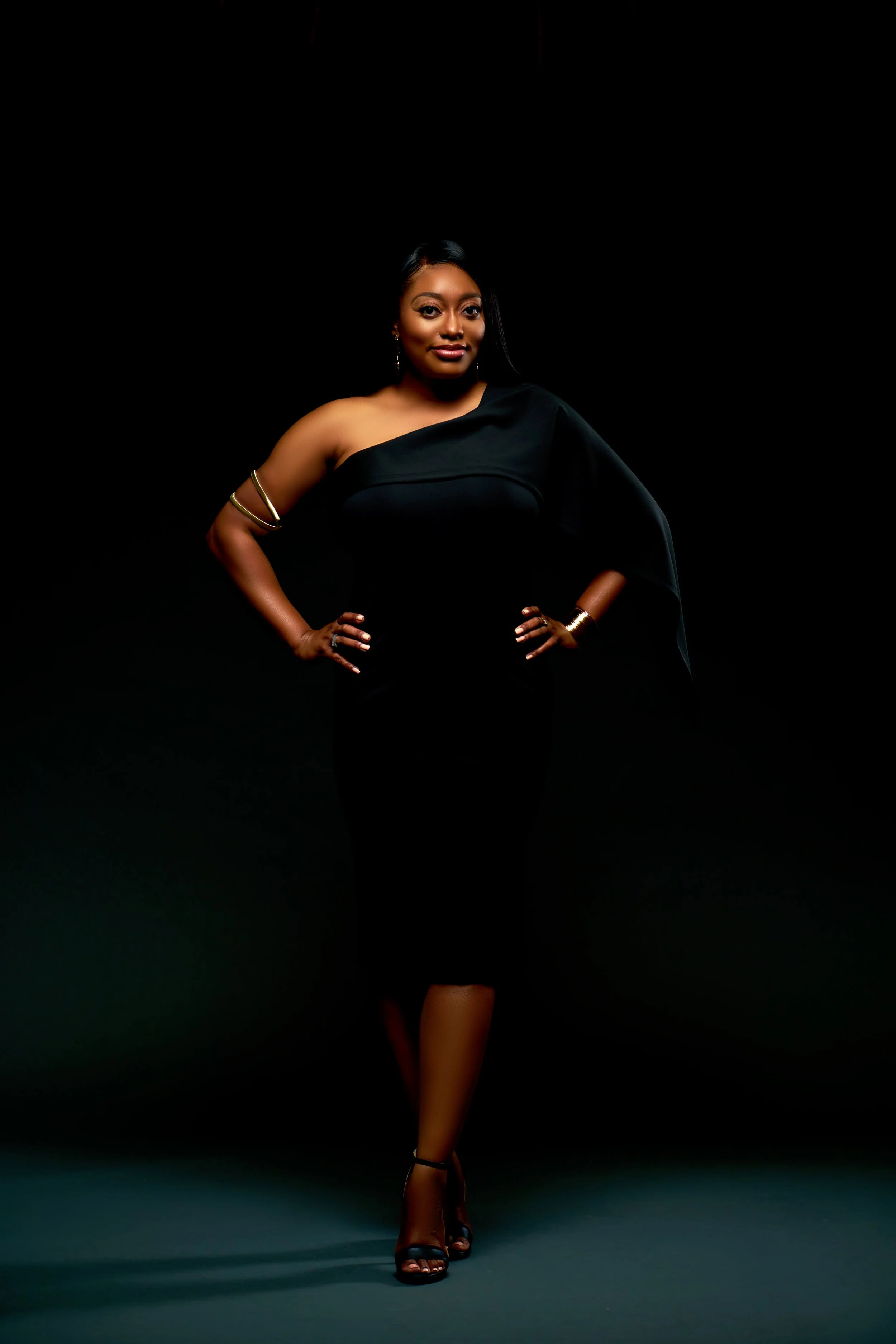 A woman with dark skin and long black hair standing against a black background, wearing a black one-shoulder dress with gold jewelry and black high-heeled sandals.