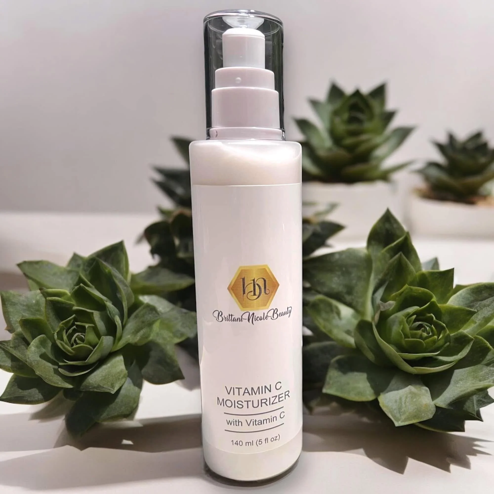 White bottle of Vitamin C moisturizer labeled by Brittan Nicole Beauty, surrounded by green leaves, against a plain background.