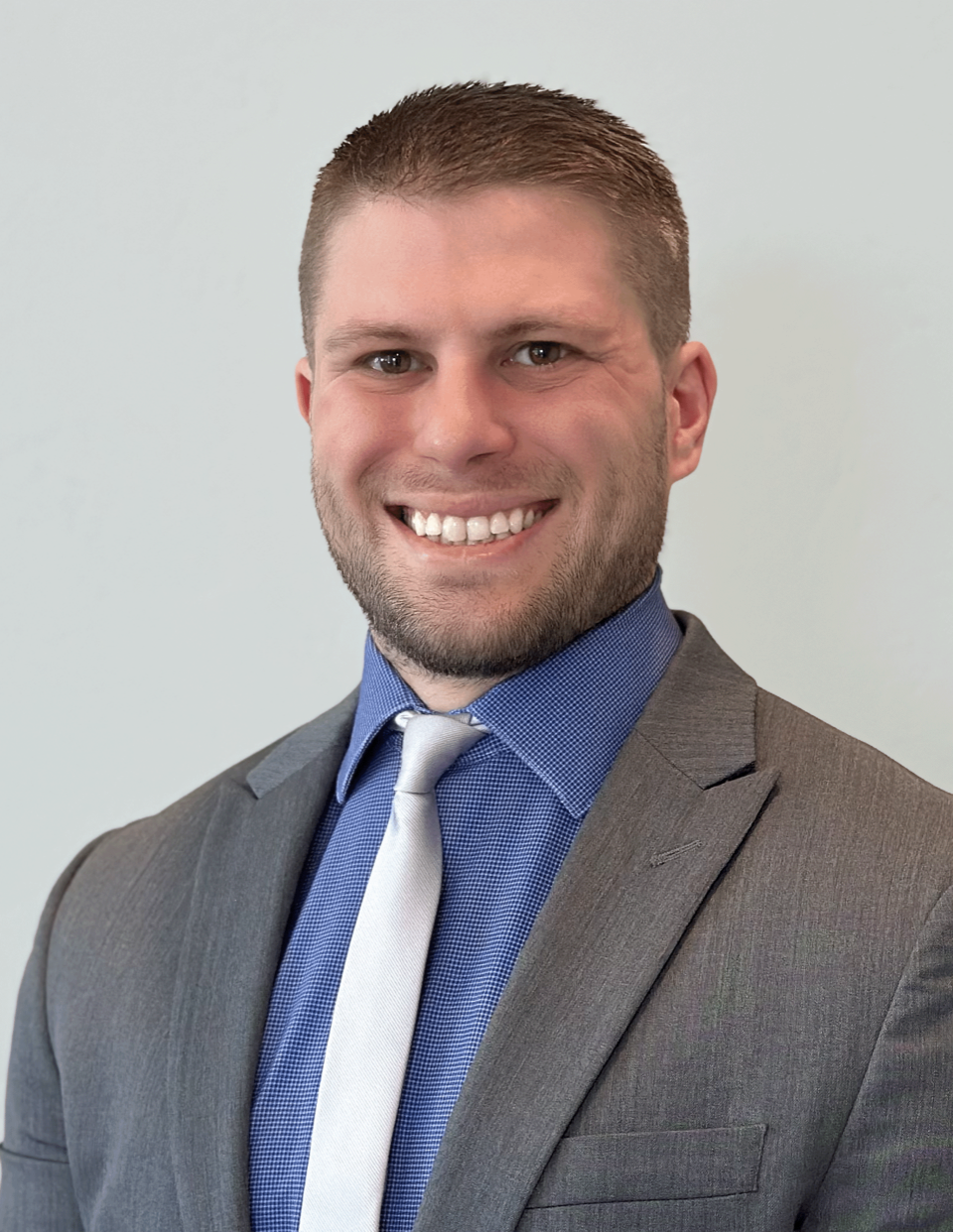 A man wearing a gray suit, blue shirt, and gray tie, smiling at the camera with a plain white background.