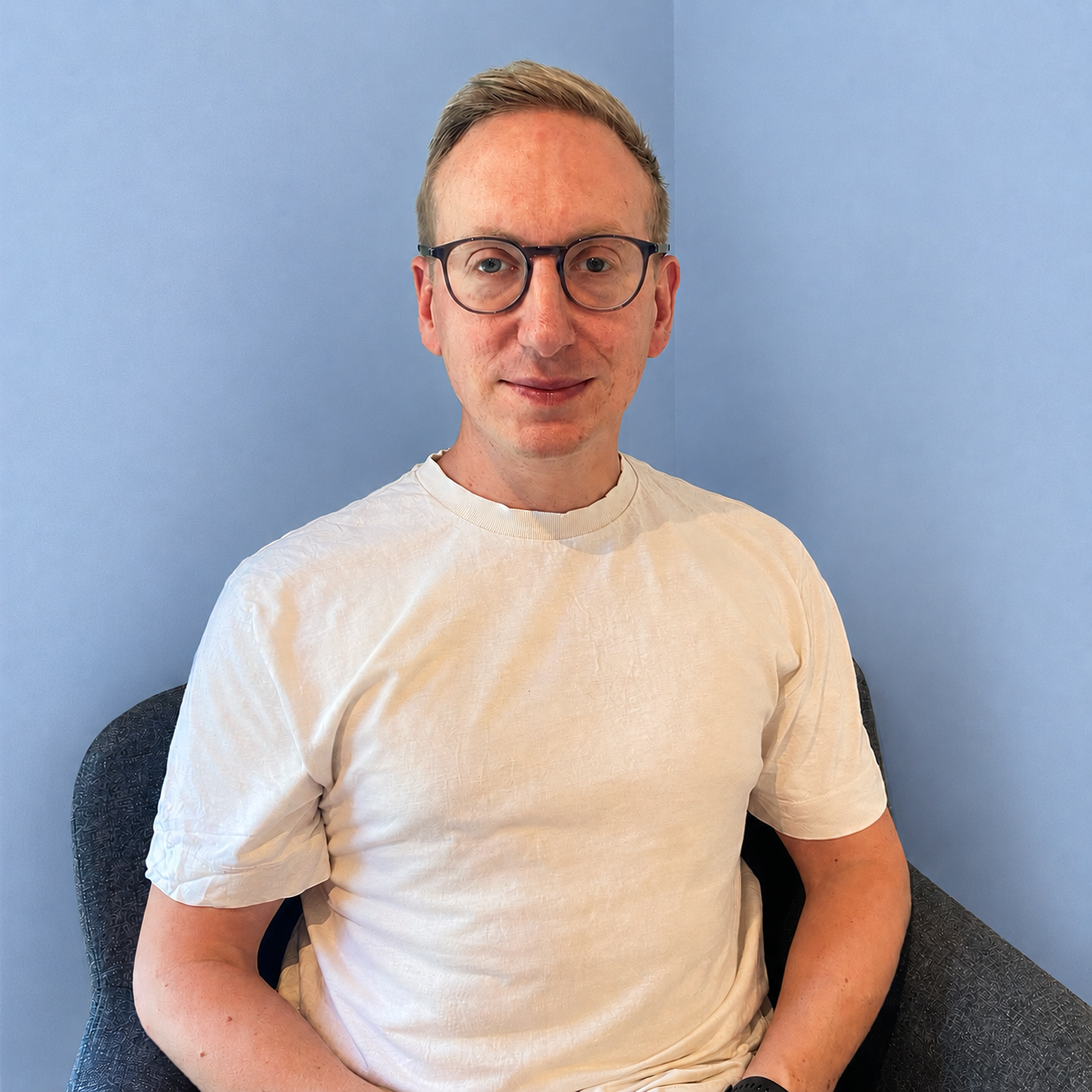 A man with glasses and short light brown hair sitting in a chair against a blue wall, wearing a white T-shirt, looking at the camera, smiling slightly.