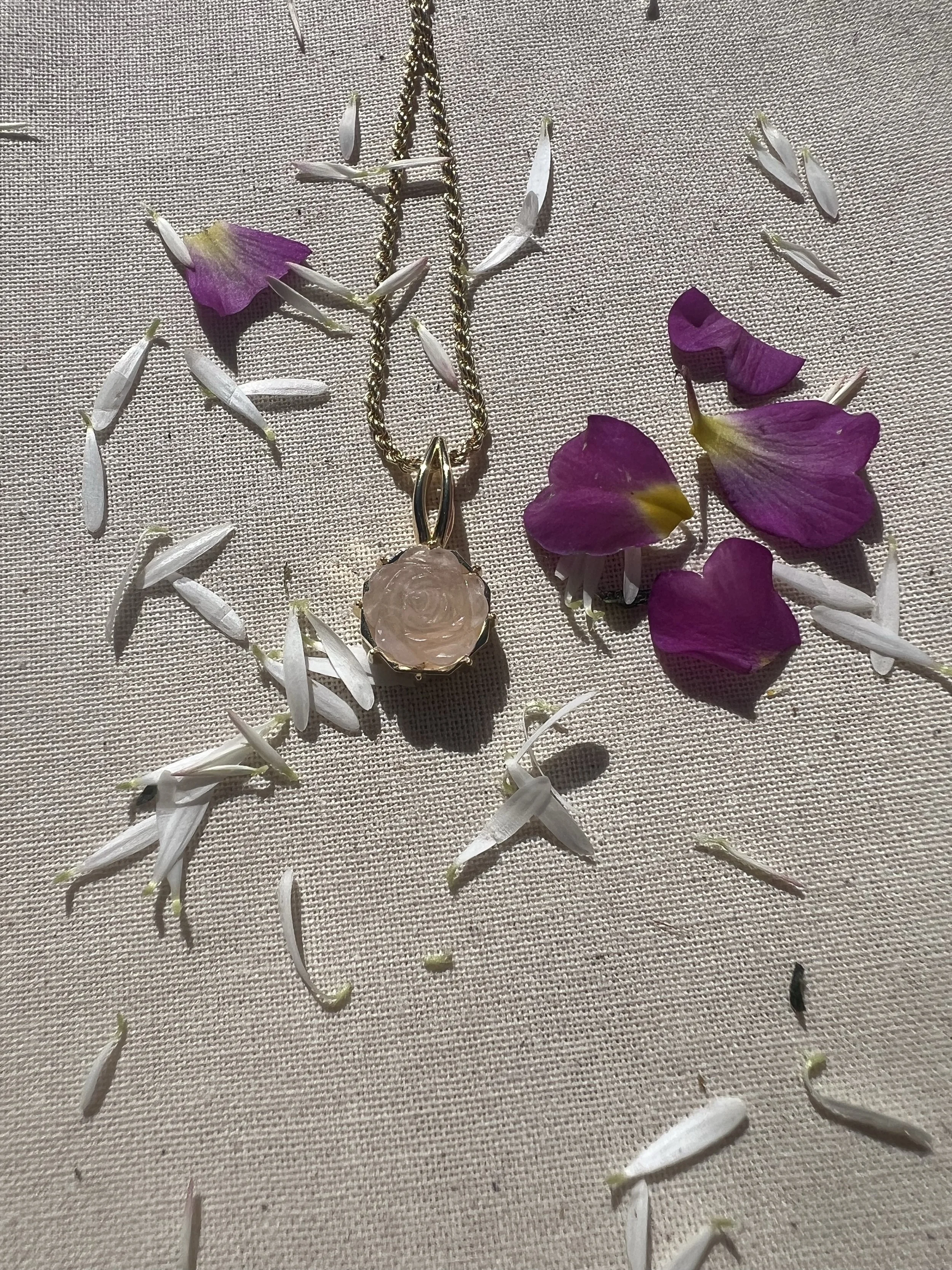 Ein goldene Kette mit einem Anhänger in Form einer rosa Blume liegt auf einem Leinentuch, umgeben von lila und weißen Blütenblättern.