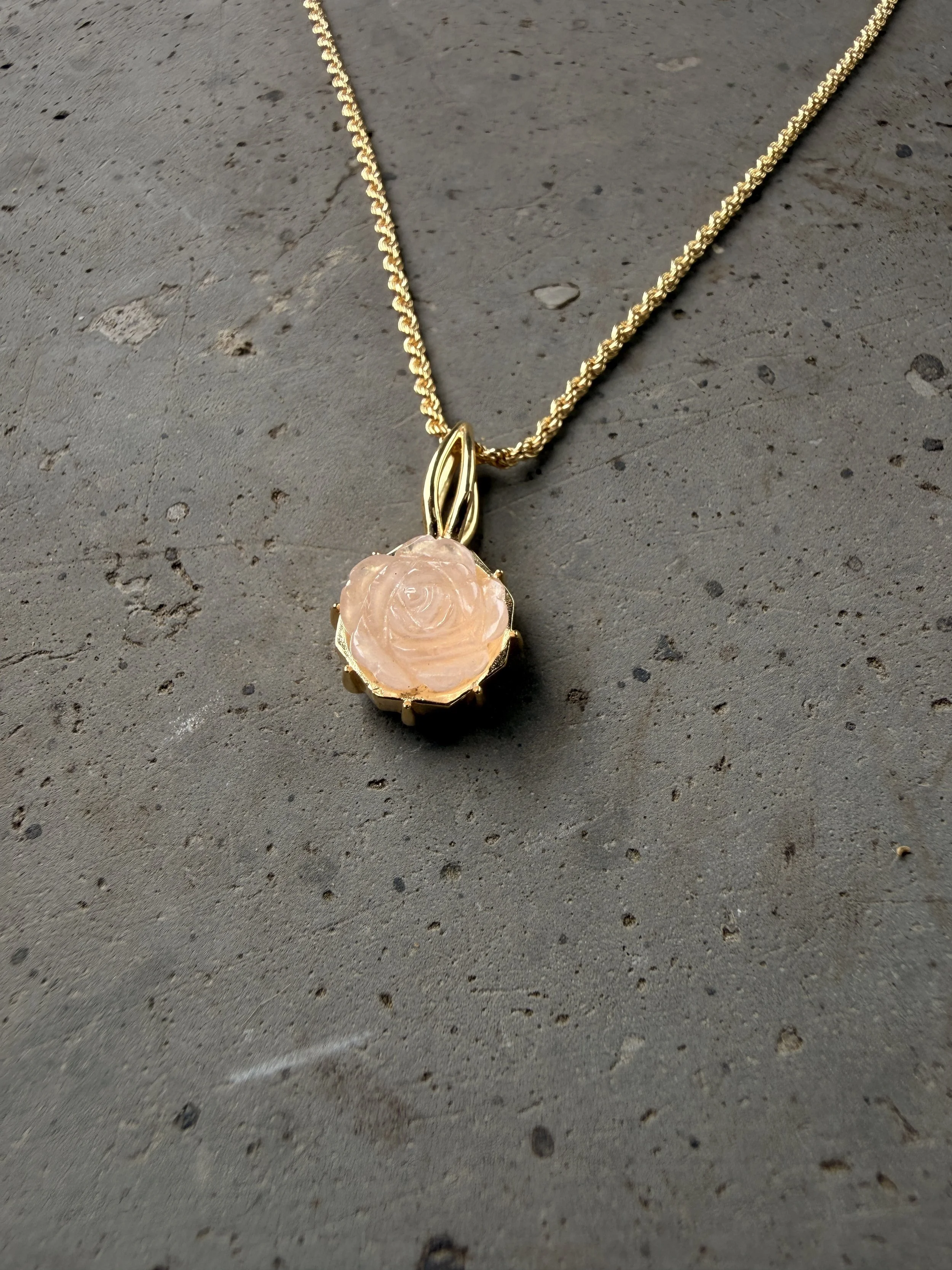Eine goldene Halskette mit einer rosa Rosenquarz-Blume am Ende liegt auf einer grauen, rauen Steinoberfläche.