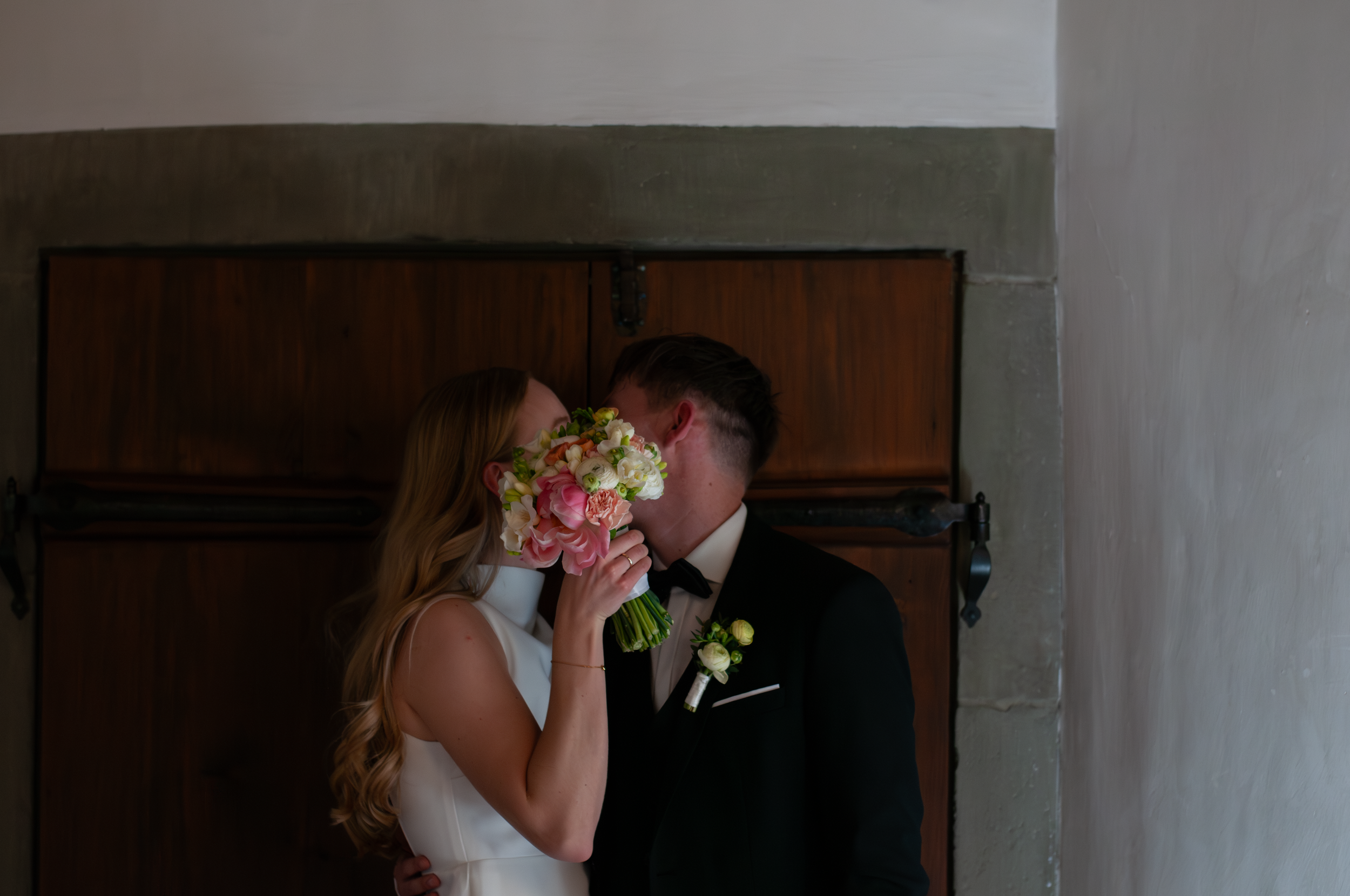 Ein Brautpaar küsst sich liebevoll, die Braut hält einen bunten Blumenstrauß vor das Gesicht, sie tragen formelle Hochzeitskleidung, im Hintergrund eine alte Holztür.