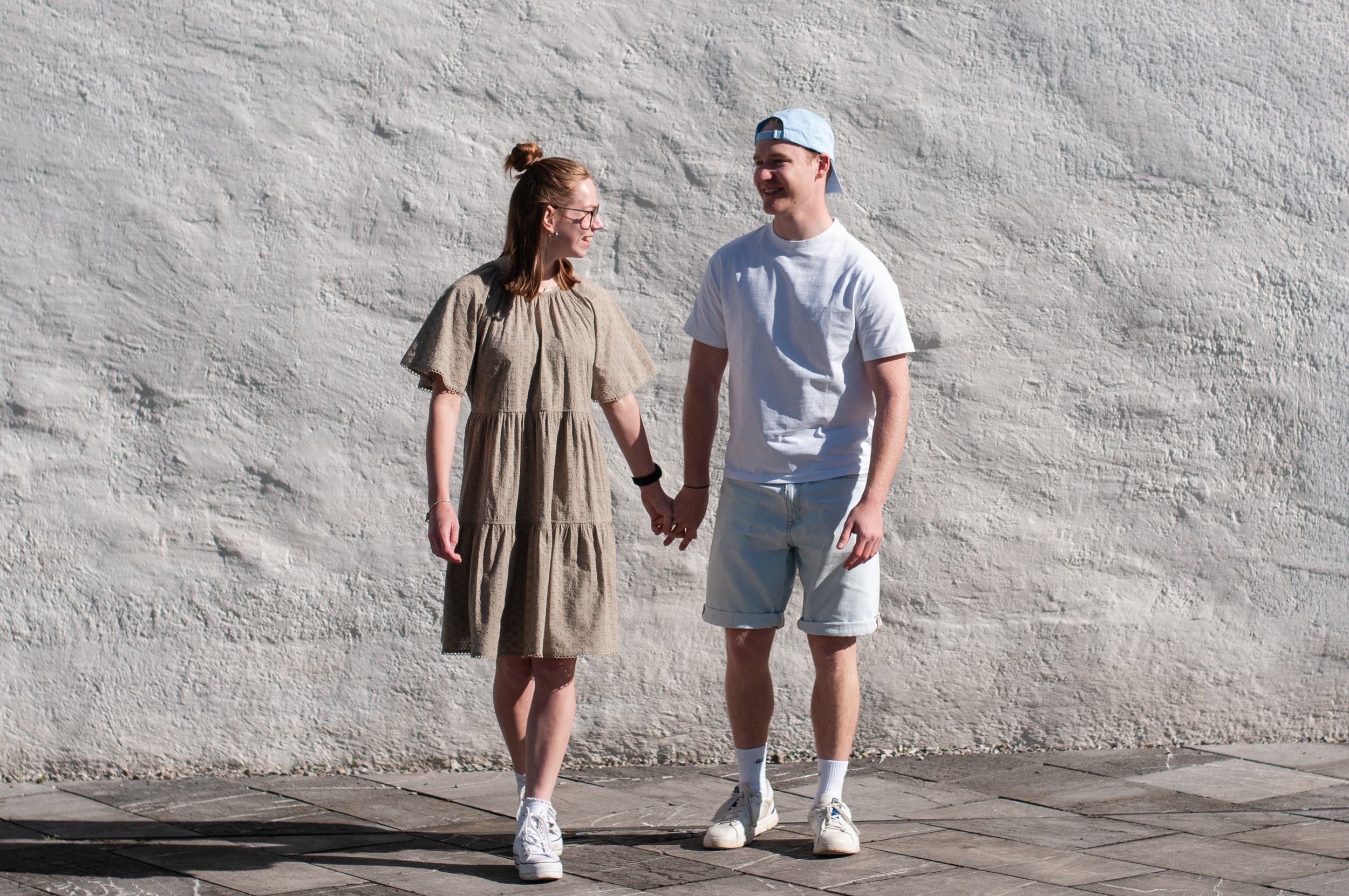 Ein junger Mann und eine junge Frau halten sich an den Händen, laufen nebeneinander vor einer weißen Wand und lachen.