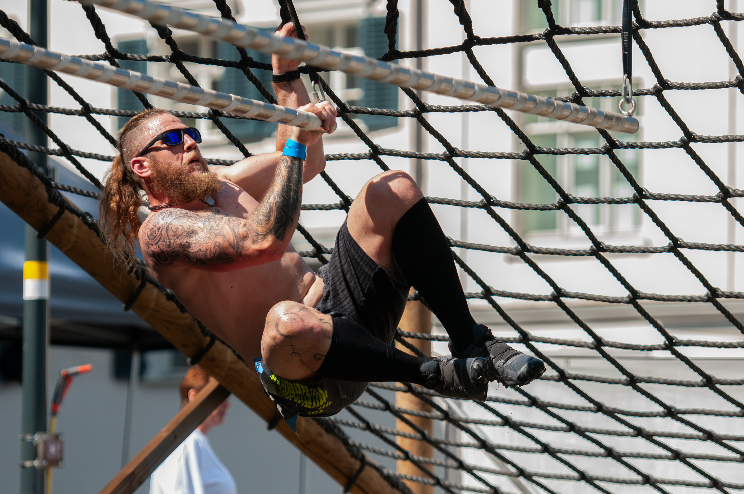 Mann klettert auf einem Klettergerüst bei einem Outdoor-Event, trägt Sonnenbrille, Tattoos, Armbänder und Sportswear, im Hintergrund Gebäude und andere Menschen.