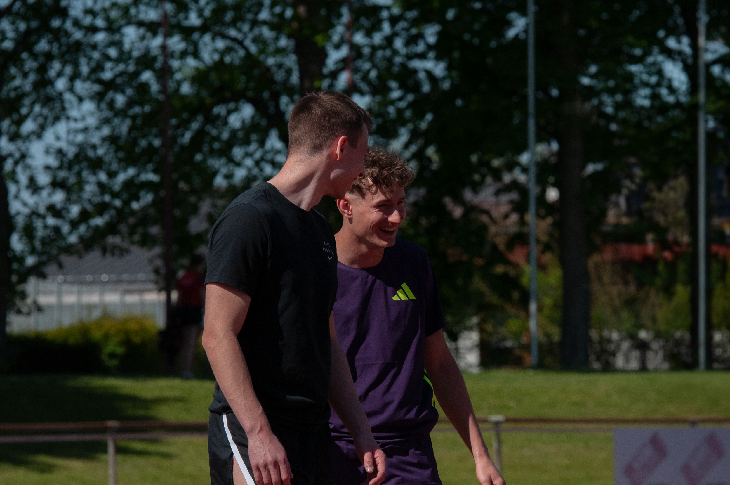 Zwei junge Männer lachen und unterhalten sich auf einem Sportplatz im Freien unter Bäumen.