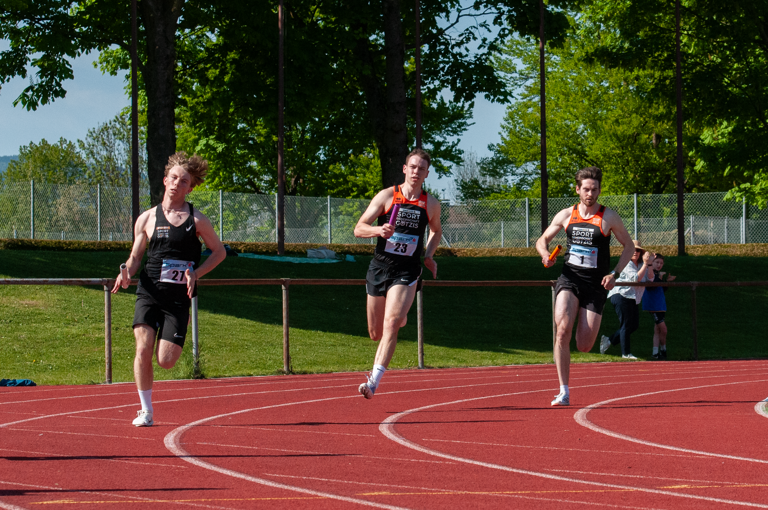Drei junge Männer laufen auf einer roten Leichtathletikbahn und tragen Sportbekleidung mit Startnummern.