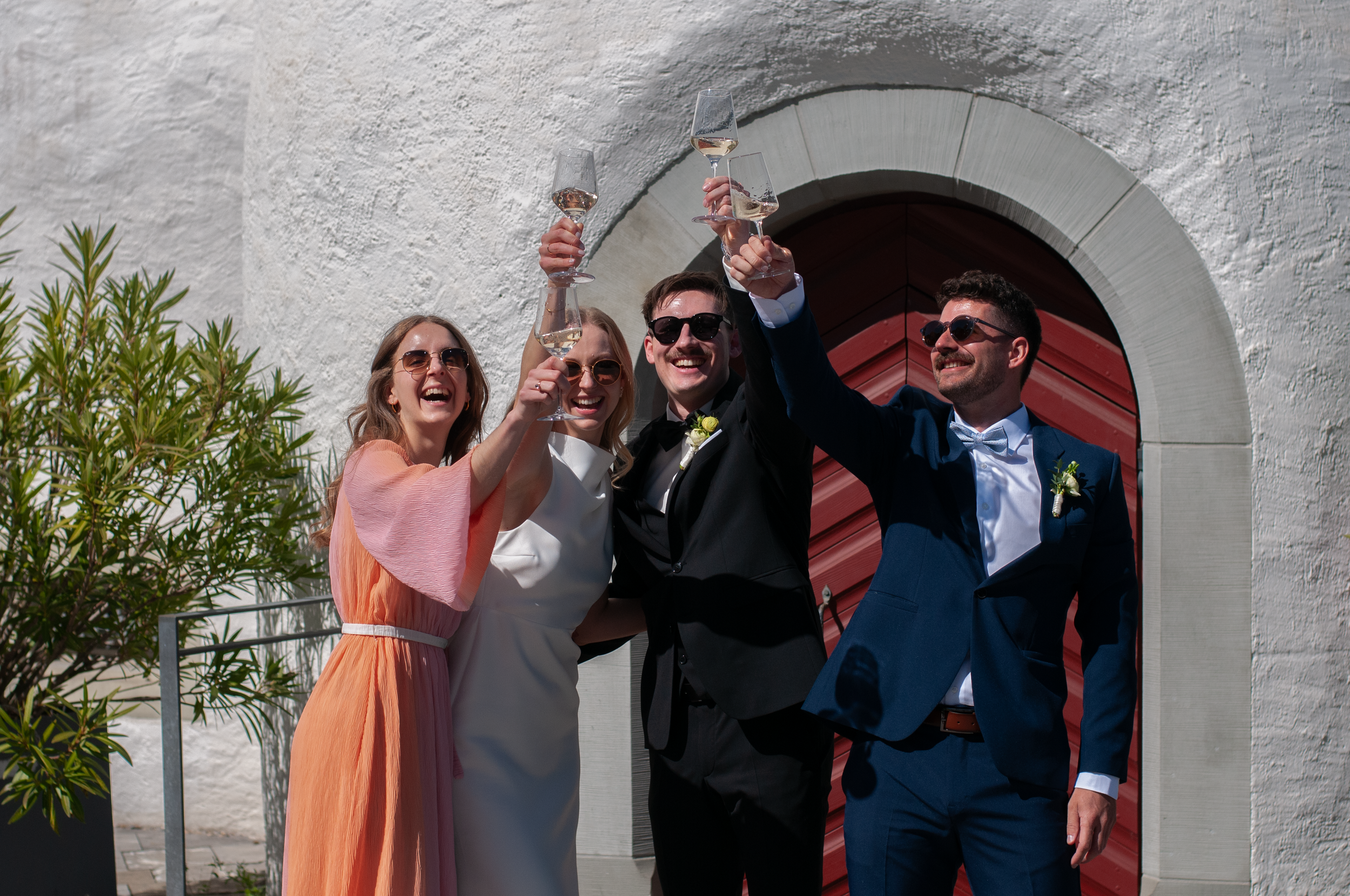 Vier Menschen feiern und stoßen mit Champagnergläsern an, vor einer weißen Wand mit rotem Tor im Hintergrund.