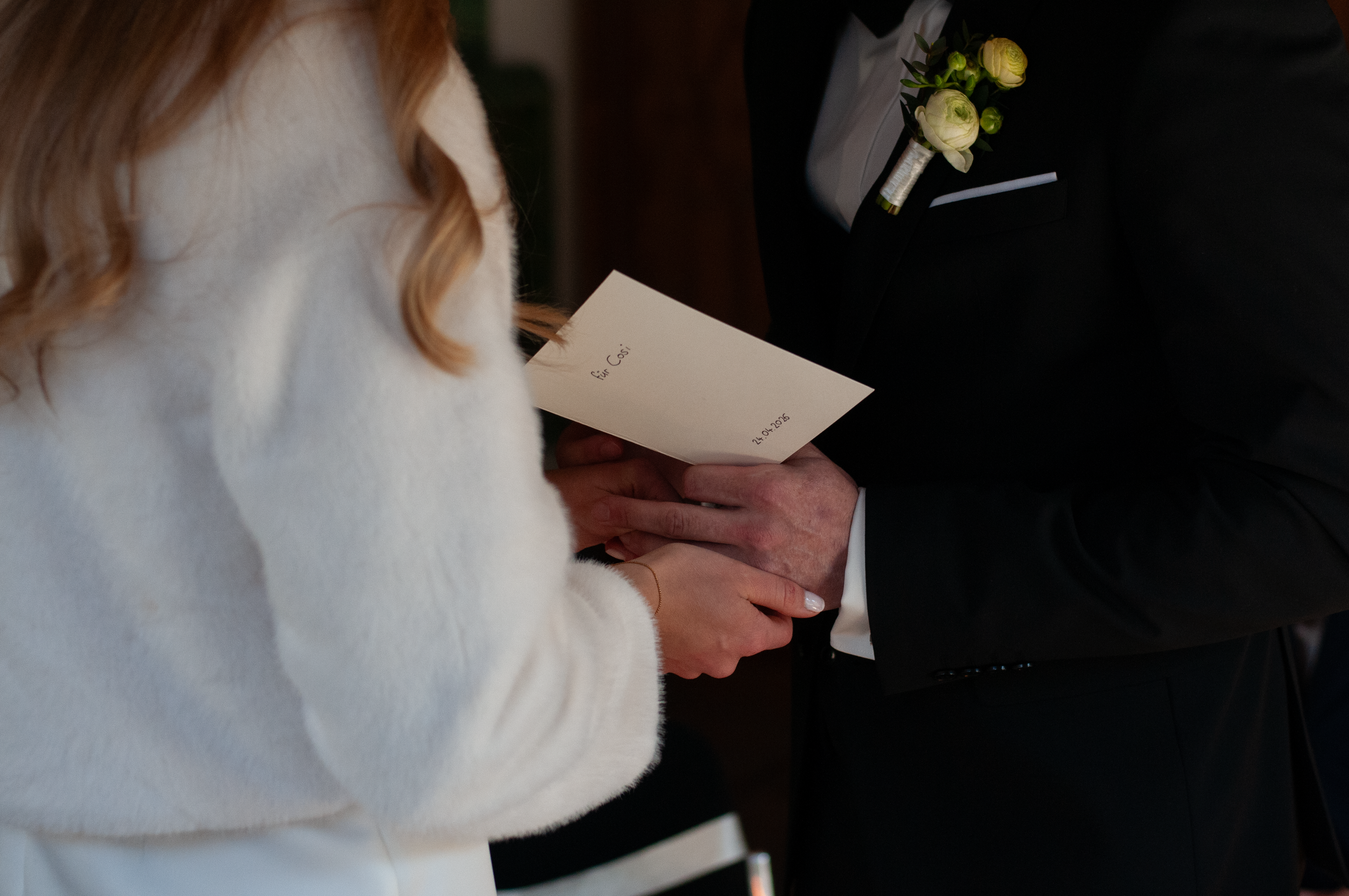 Eine Frau in einem weißen Mantel hält ein weißes Hochzeitsgedicht mit dem Datum 24.04.2026, während sie mit einem Mann in einem schwarzen Anzug auf einer Hochzeit steht, der einen Anstecker mit weißen Blumen trägt.