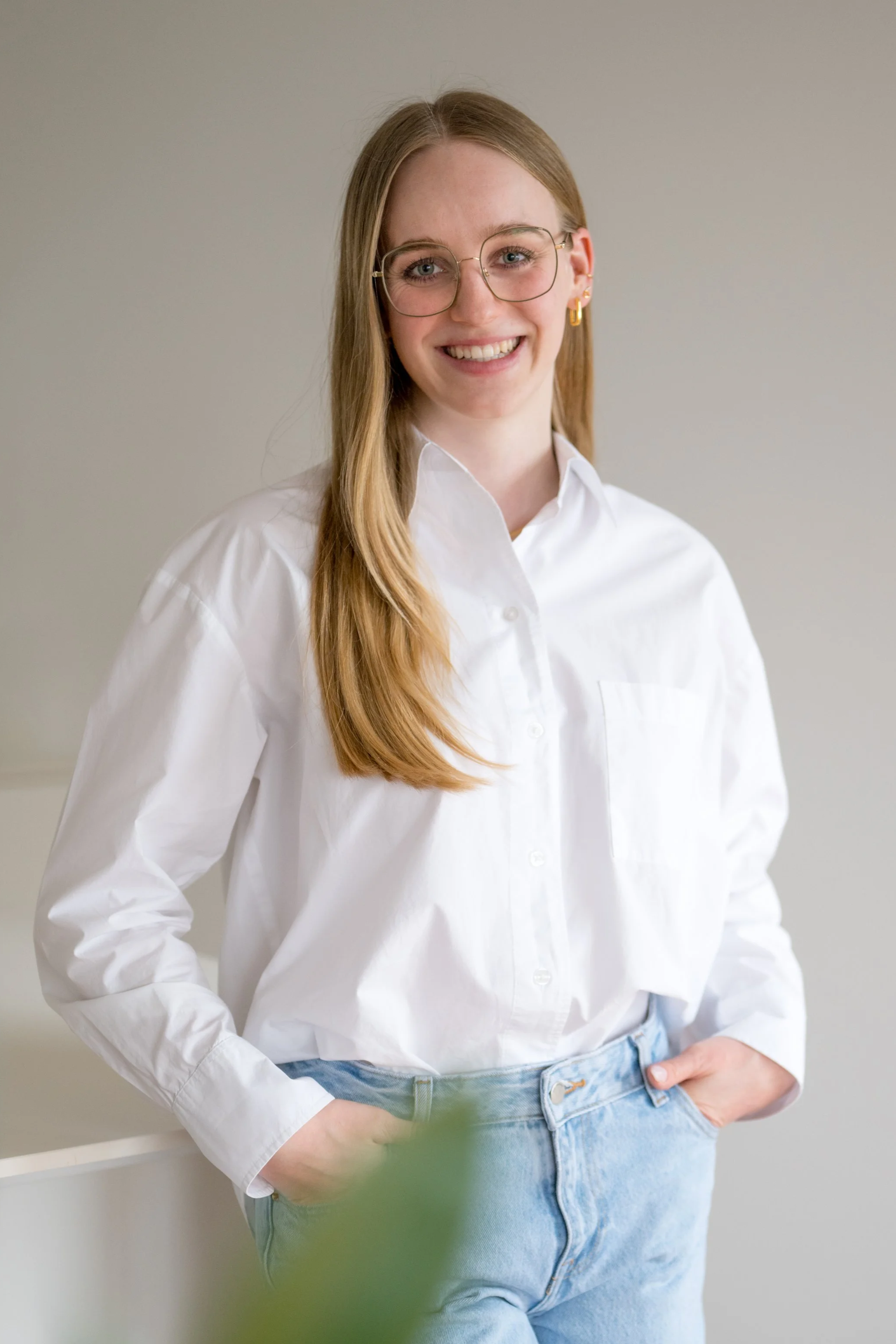 Junge Frau mit blonden Haaren, Brille, weißem Hemd und blauen Jeans, stehend vor hellem Hintergrund, lachend.