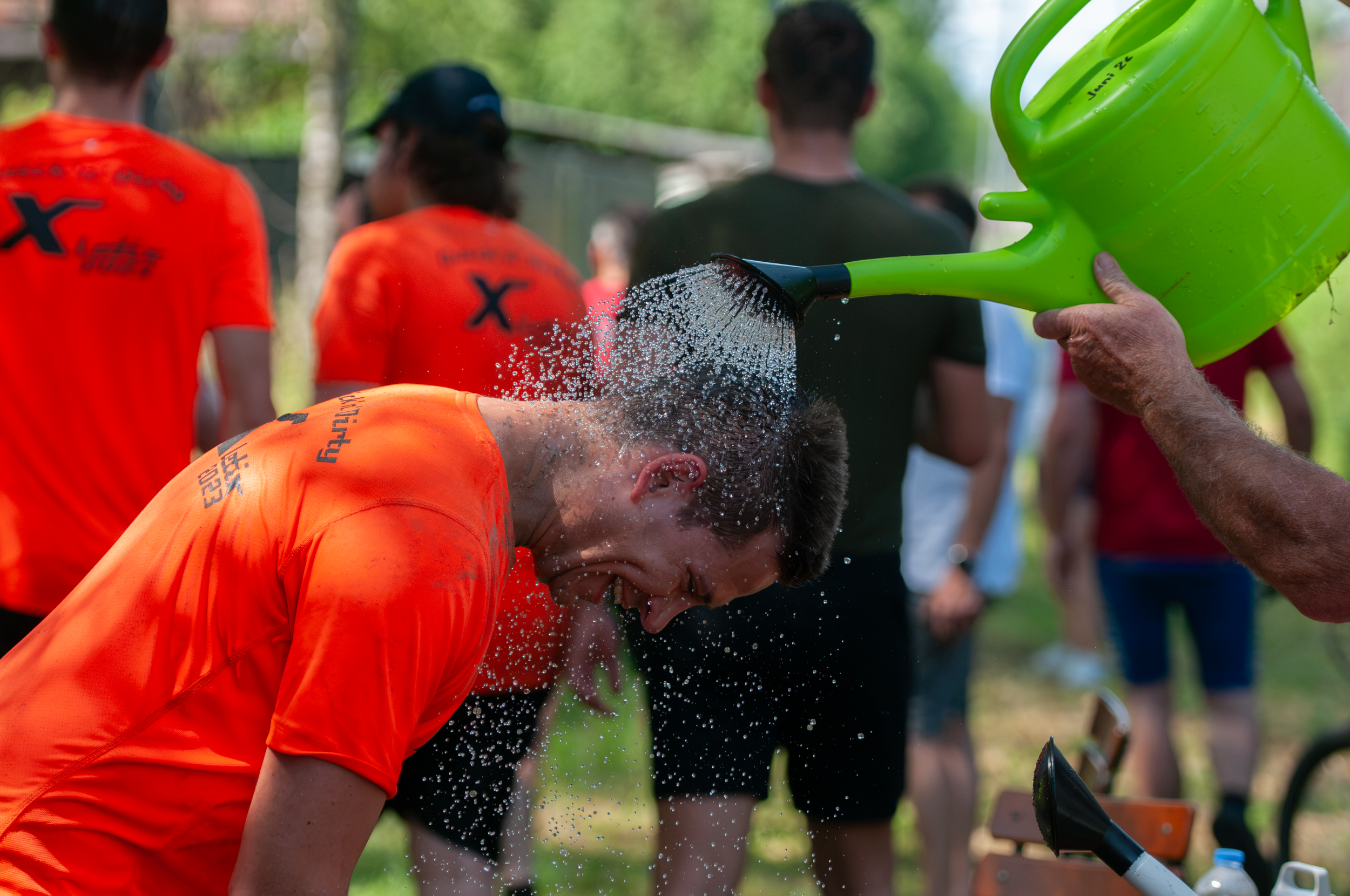 Mehrere Menschen am Strand werden mit Wasser bespritzt, einer trägt ein orangefarbenes T-Shirt, im Hintergrund sind Bäume und Leute. Es scheint eine spaßige und erfrischende Aktivität zu sein.