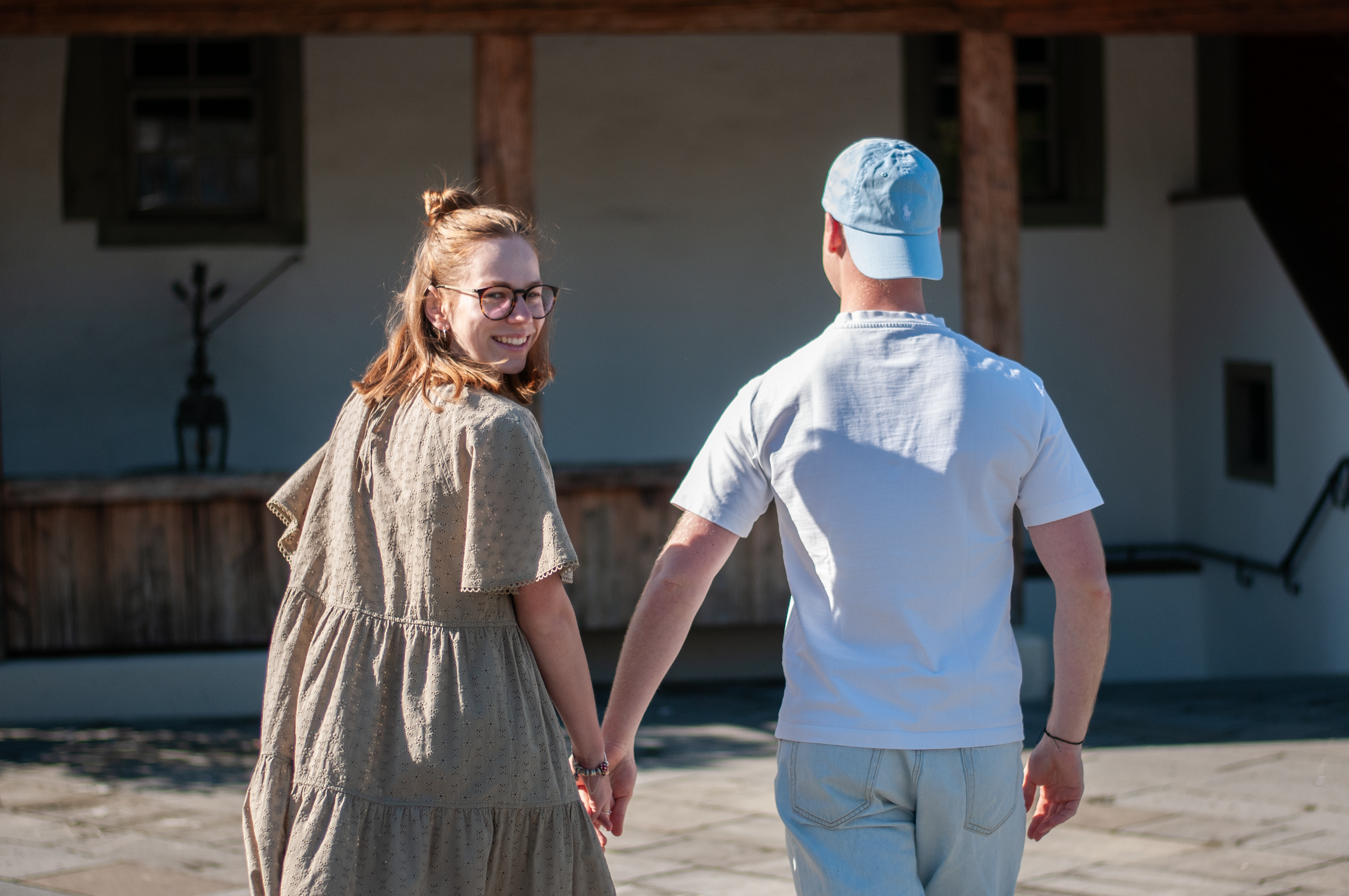 Ein Paar hält sich an den Händen und geht am Strand spazieren, die Frau trägt ein beige Kleid mit Spitzenmuster, der Mann trägt ein weißes T-Shirt und einen hellblauen Hut.
