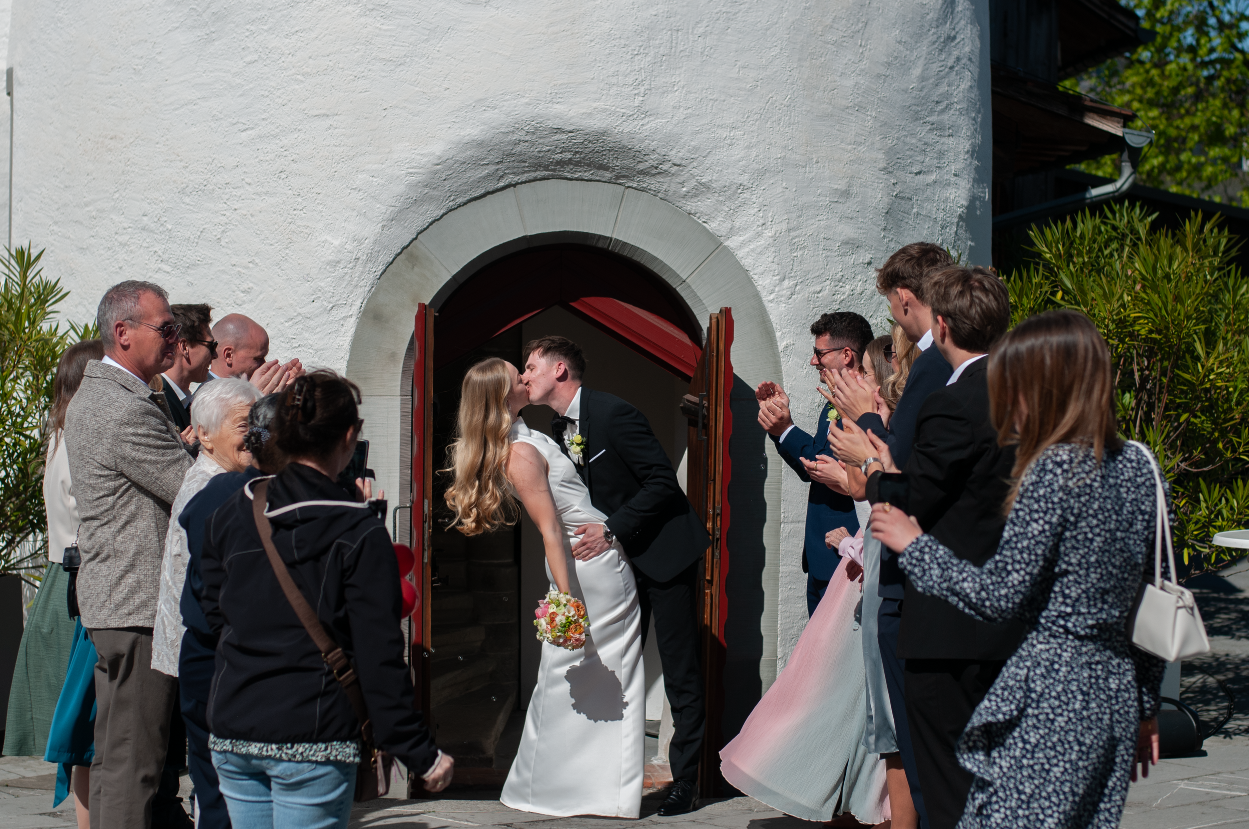 Ein Brautpaar küsst sich vor der Tür einer weißen Kirche, während ihre Gäste applaudieren und sie auf der Hochzeit begrüßen.