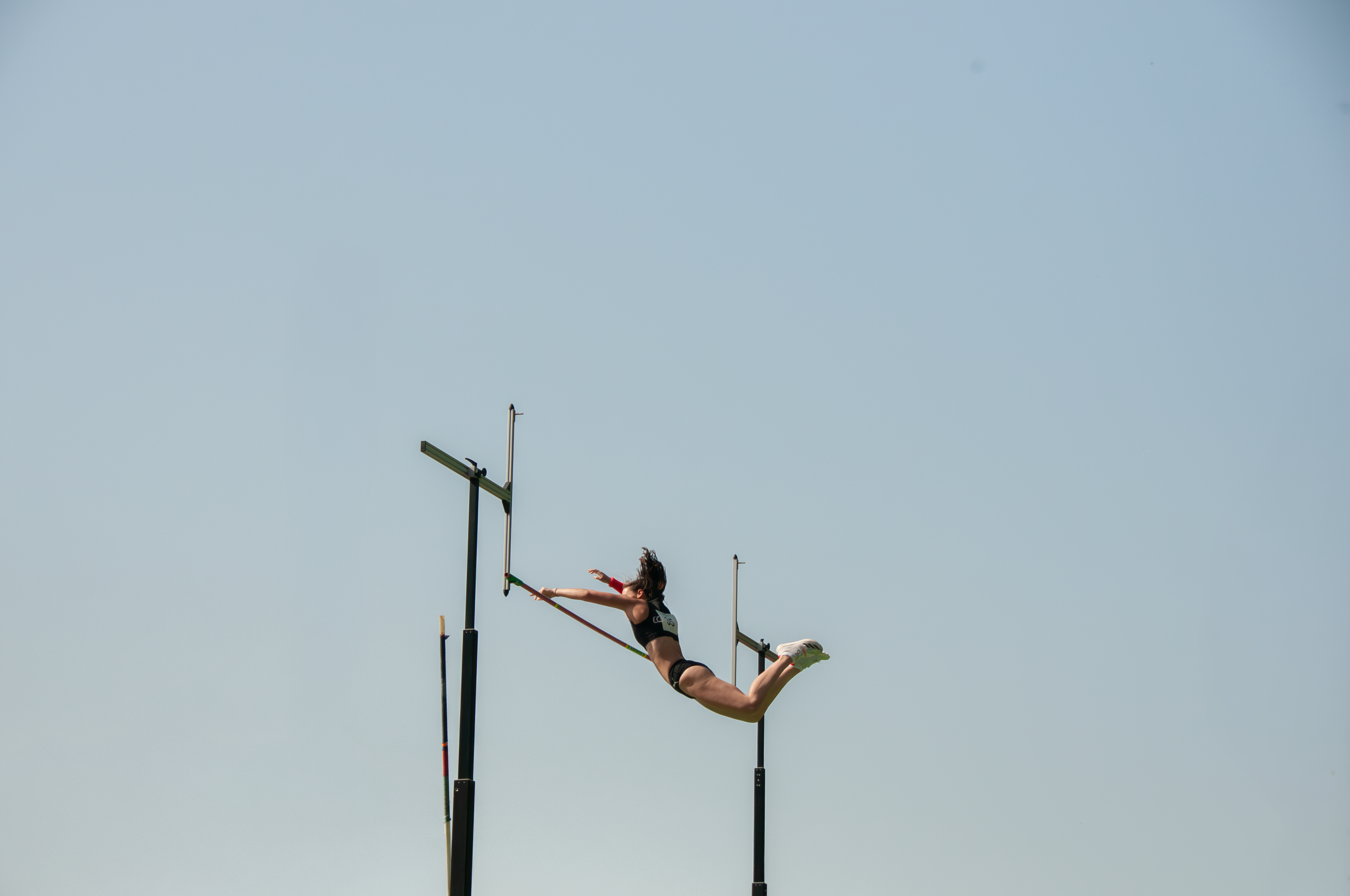 Eine Athlete, die auf einem Stabhochsprung-Gerüst überspringt, während sie in der Luft ist, bei einem Sportwettbewerb; die Person trägt eine schwarze Sportbekleidung und hat braune Haare.