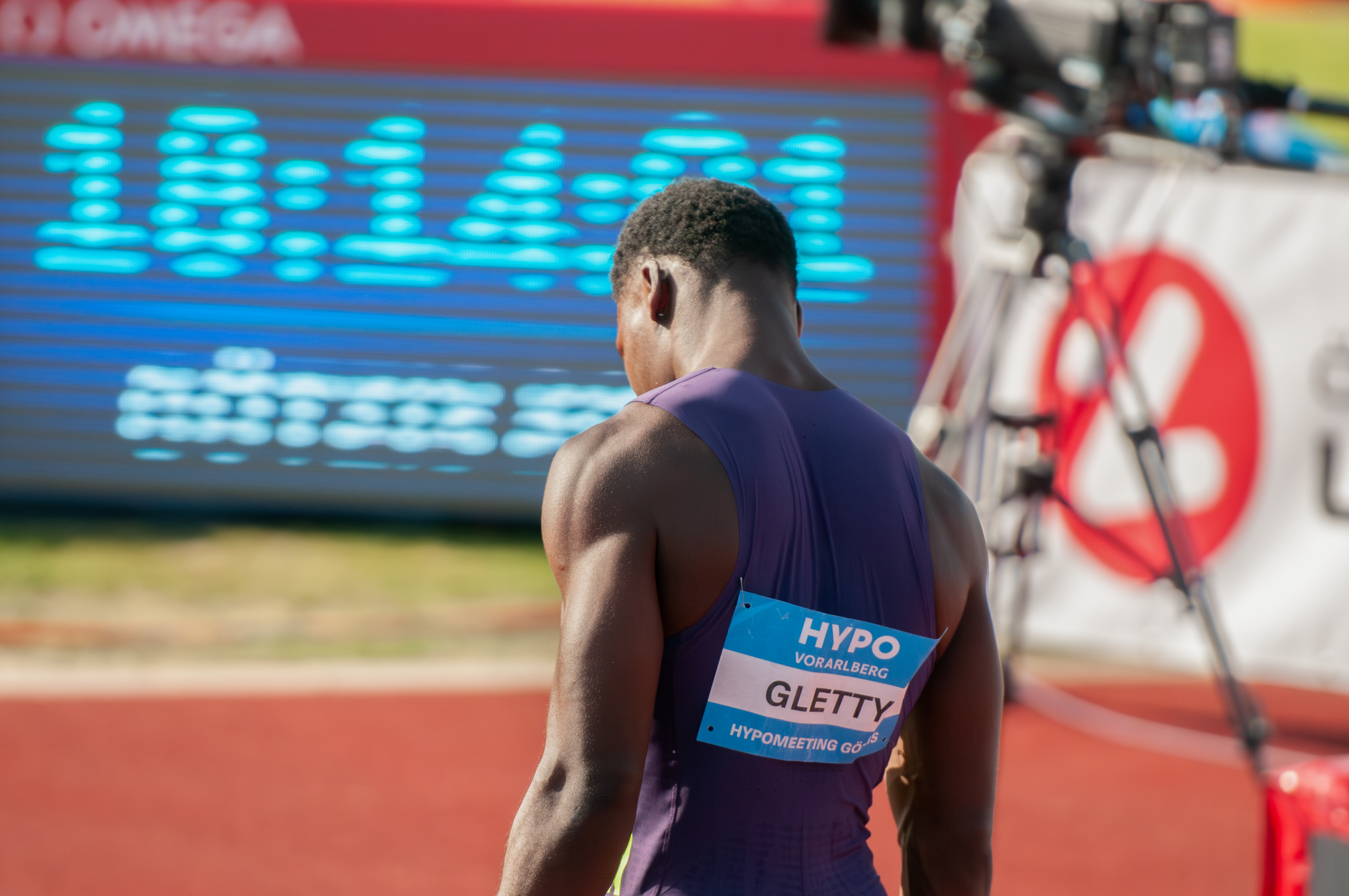 Leistungssportler am Start bei einem Wettkampf, Blick auf den Rücken, Laufnummer GLETTY, Umgebung eines Stadion mit Anzeigetafel im Hintergrund.