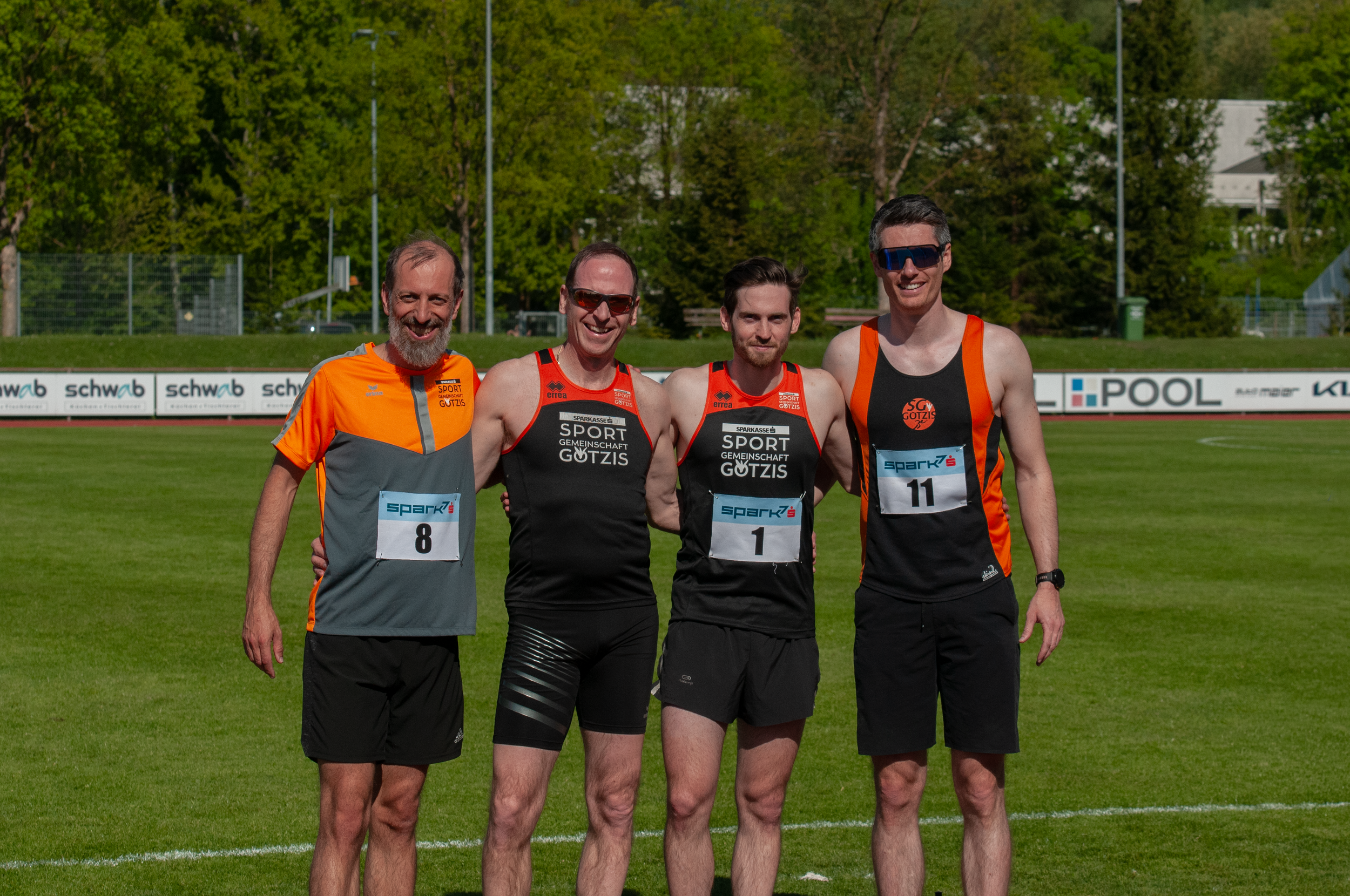 Vier Männer in Sportkleidung stehen auf einem Sportplatz, lächeln und posieren für die Kamera. Es ist sonnig, im Hintergrund Bäume und eine Laufbahn sichtbar.