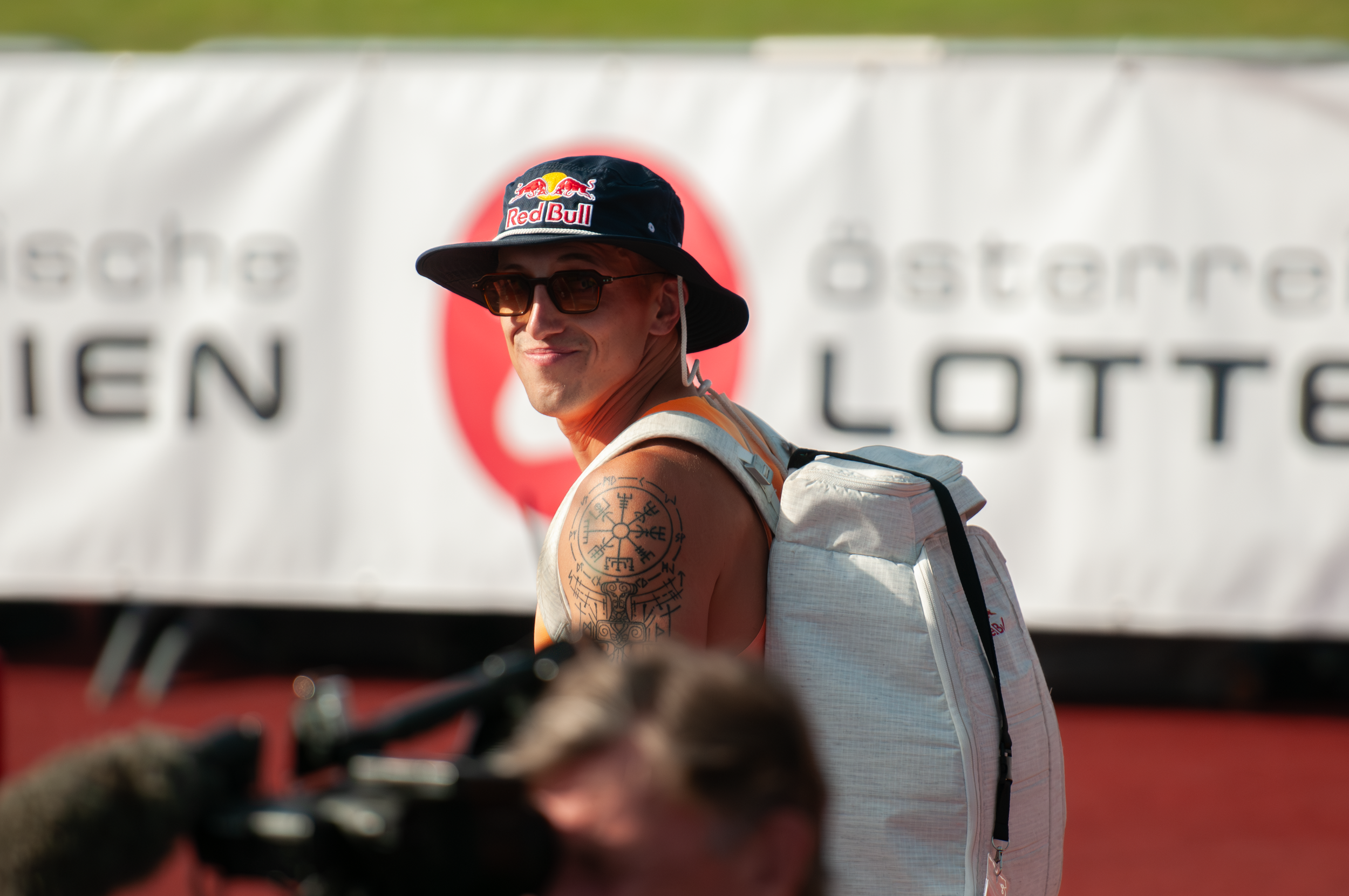 Junger Mann trägt einen Sonnenhut mit Red Bull Logo, Sonnenbrille und Rucksack, steht vor einem weißen Hintergrund mit rotem Logo.