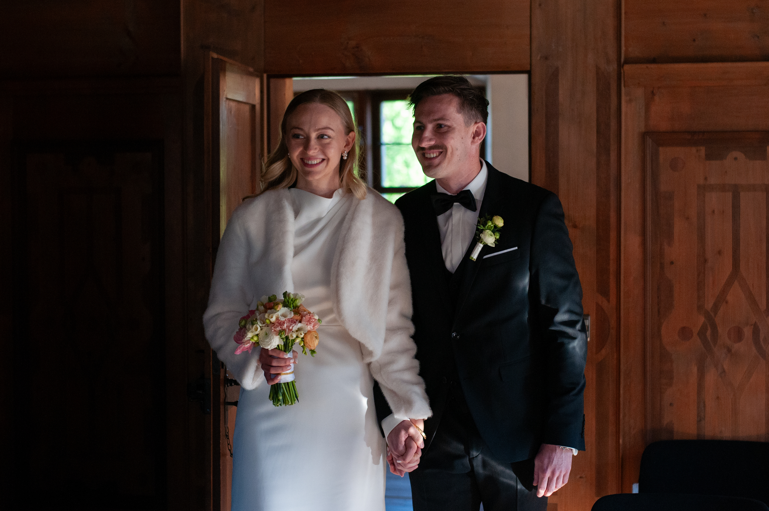Ein Brautpaar beim Heiratsakt in einem Raum mit Holzvertäfelung, die Braut trägt ein weißes Kleid und einen Pelzmantel, hält einen Blumenstrauß, der Bräutigam trägt einen schwarzen Anzug mit Fliege und hat eine kleine Blumenbrosche auf der Jacke.