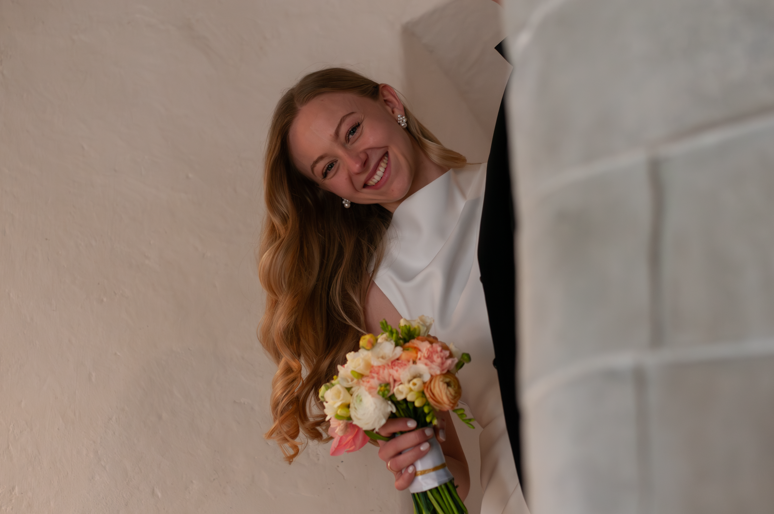 Eine lachende Frau mit langen, lockigen roten Haaren trägt eine weiße Kleidung und trägt Ohrringe. Sie hält einen bunten Blumenstrauß in der Hand und steht vor einer weißen Wand.