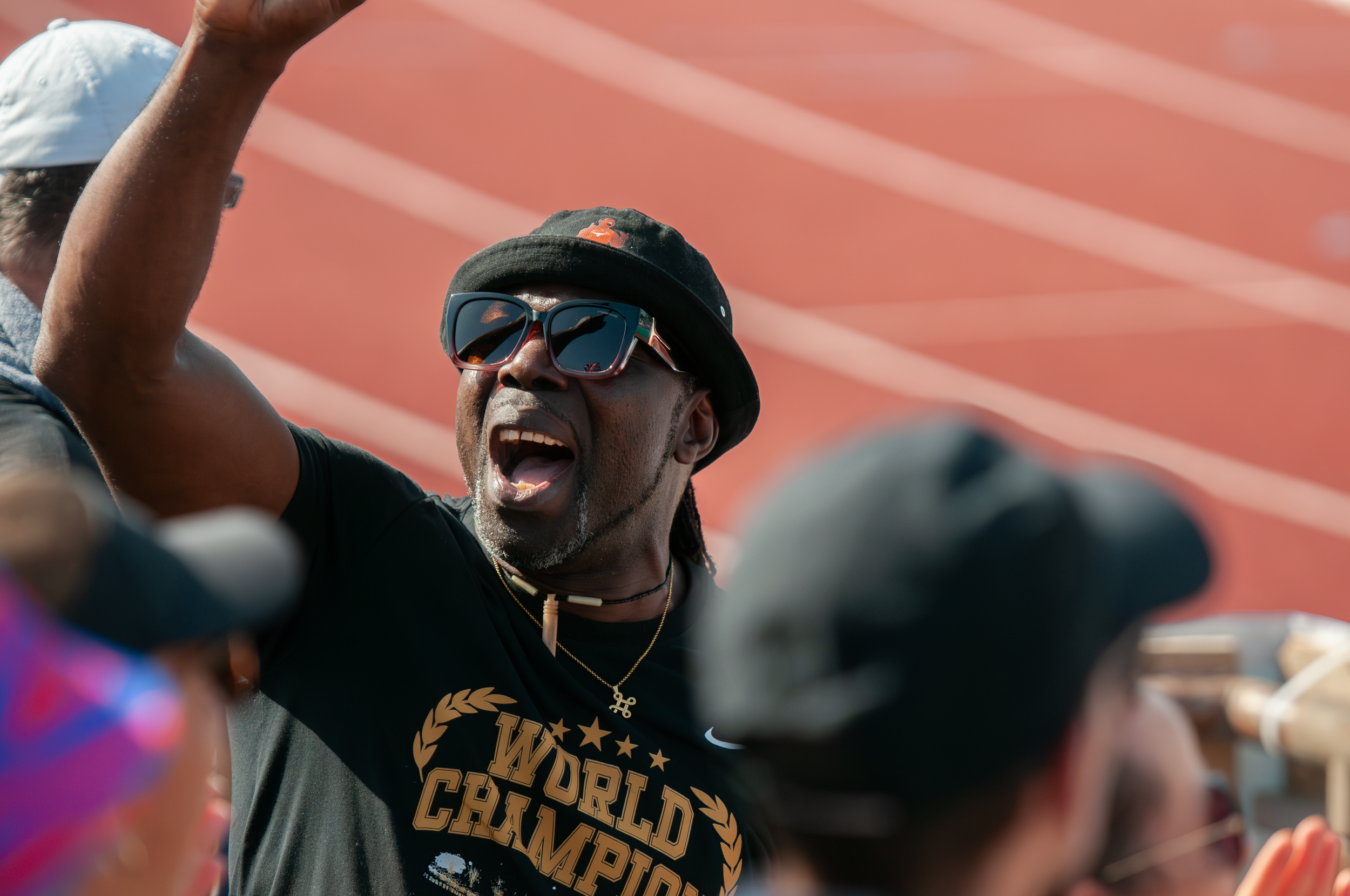 Ein Mann mit Sonnenbrille, schwarzer Kappe und einem schwarzen T-Shirt, das den Text 'World Champions' trägt, ist bei einem Sportevent. Er wirkt enthusiastisch, mit offenem Mund, während er in einer Trainingsrunde oder beim Wettkampf ist.