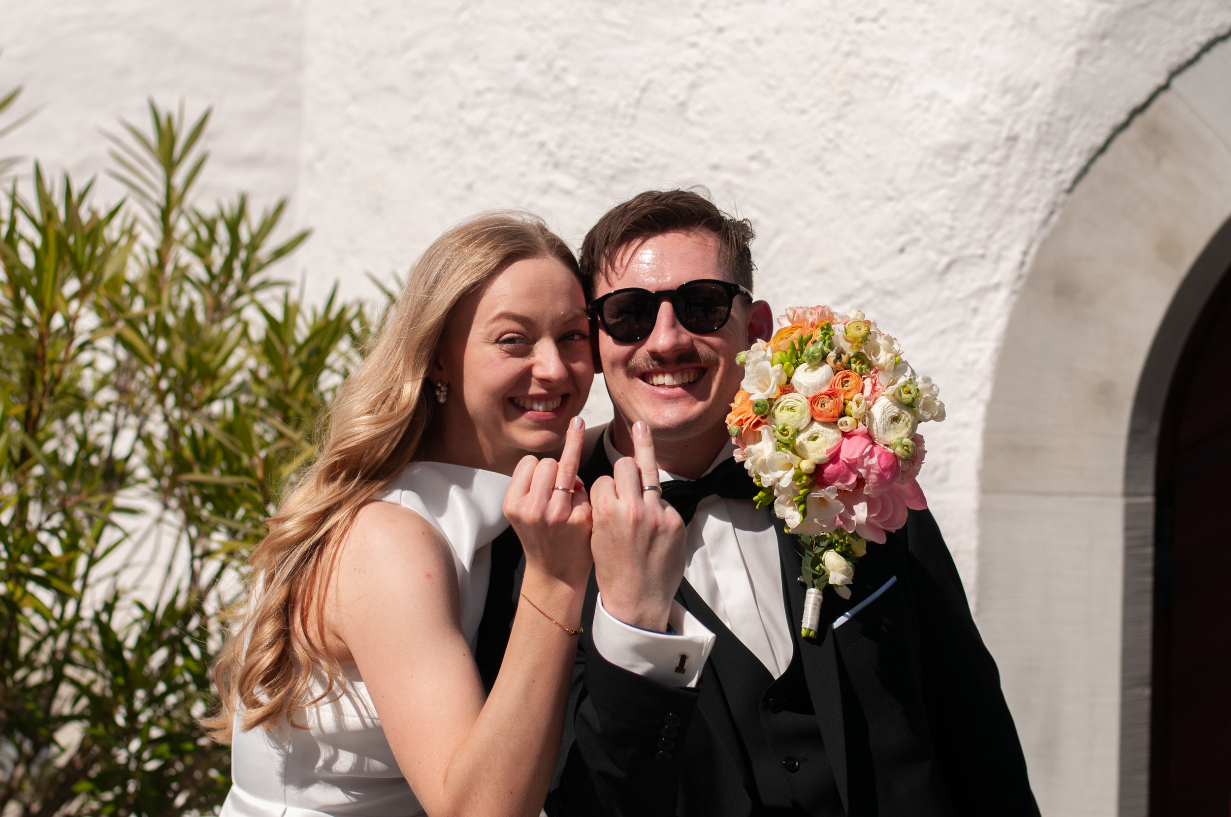 Ein Liebespaar auf ihrer Hochzeit, der Mann trägt einen schwarzen Smoking und eine schwarze Sonnenbrille, während die Frau ein weißes Hochzeitskleid trägt. Sie halten ein Blumenbouquet und zeigen ihre Finger mit Eheringen.