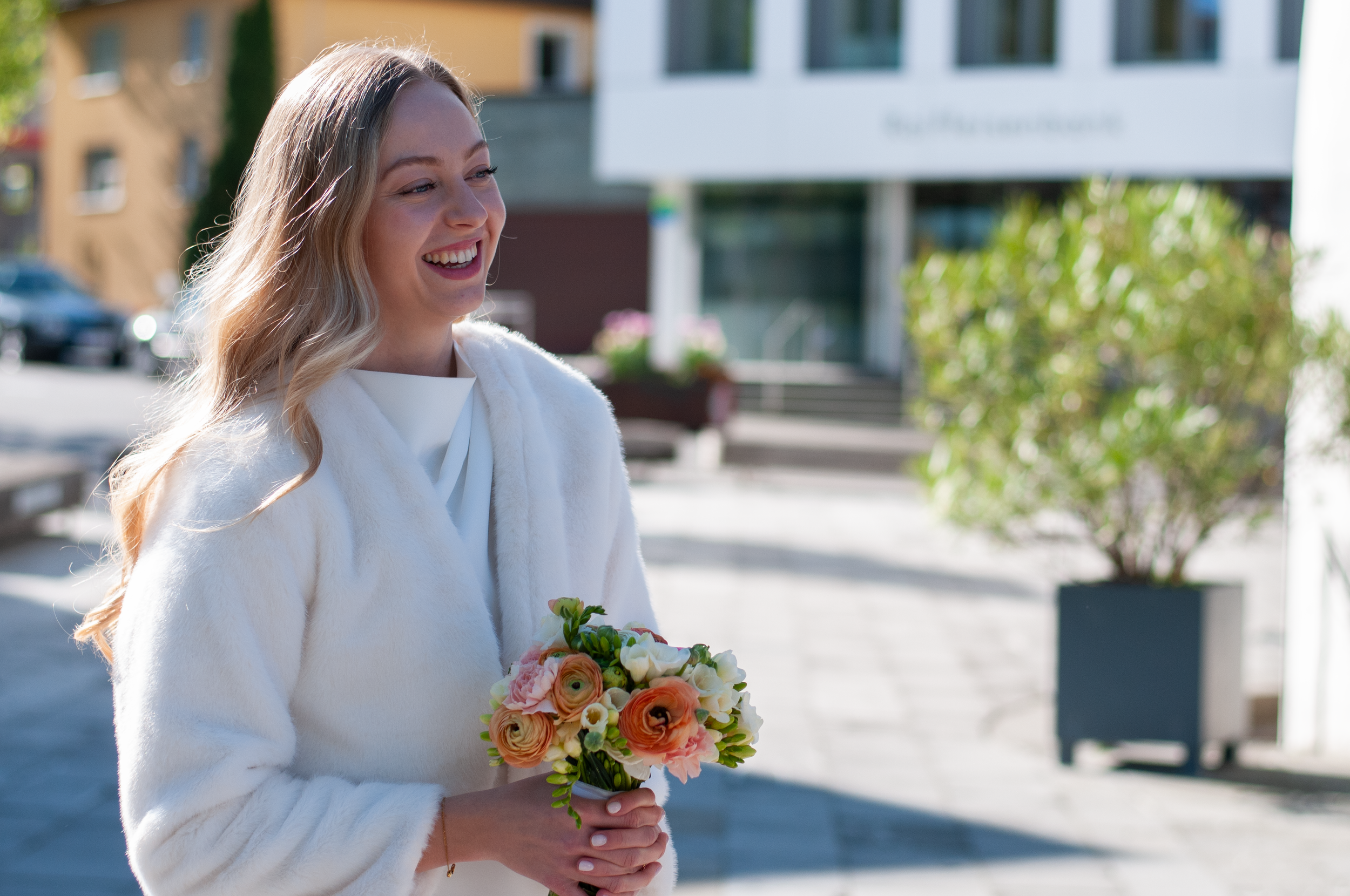 Eine lachende Frau mit langem, blondem Haar trägt einen weißen Mantel und hält einen Blumenstrauß mit rosa, orange und weißen Blumen. Sie steht draußen vor modernen Gebäuden bei Tageslicht.
