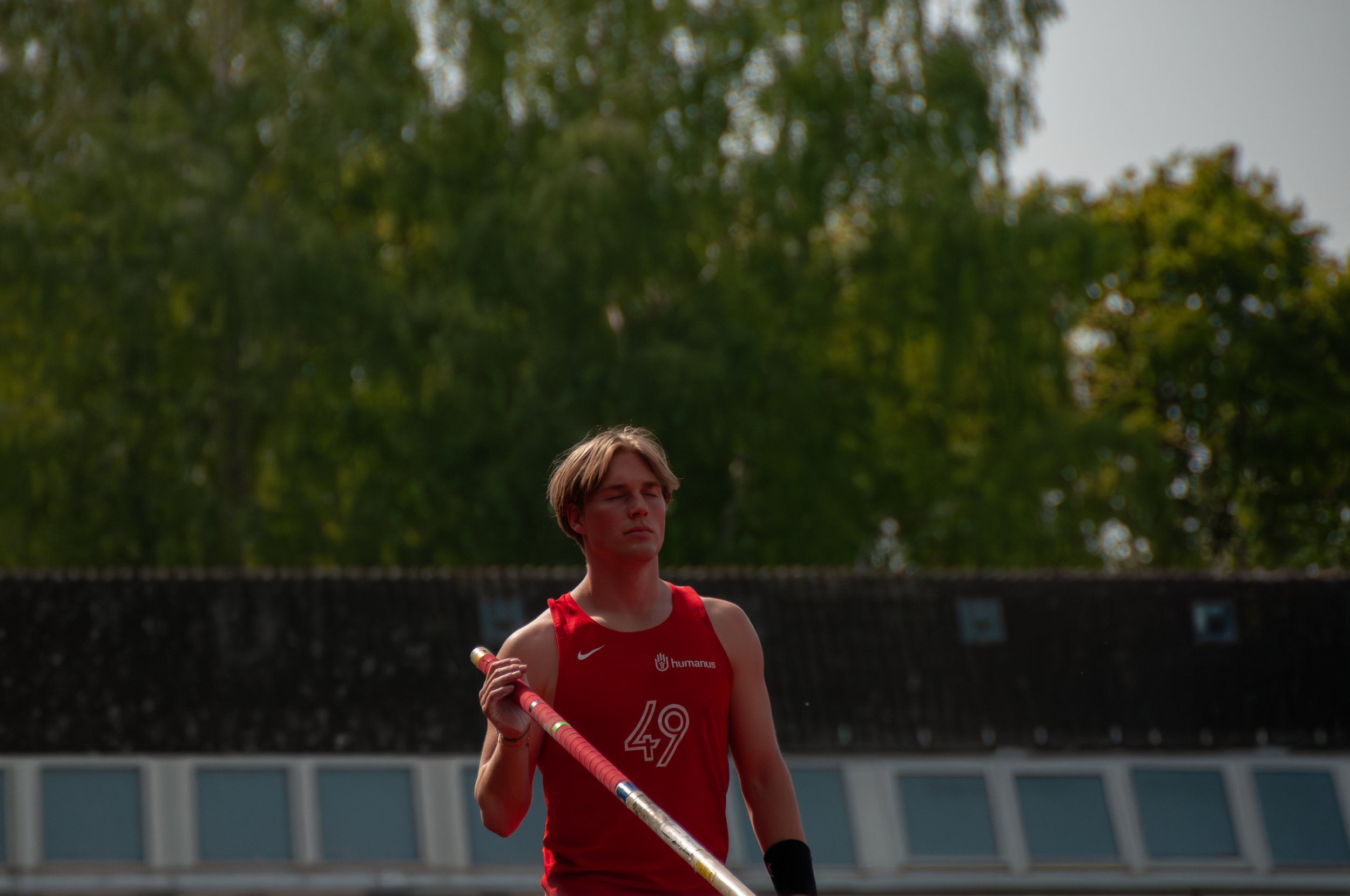 Junge weibliche Leichtathletin in rotem Sporttrikot bei Stabhochsprung auf einem Sportplatz umgeben von Bäumen.