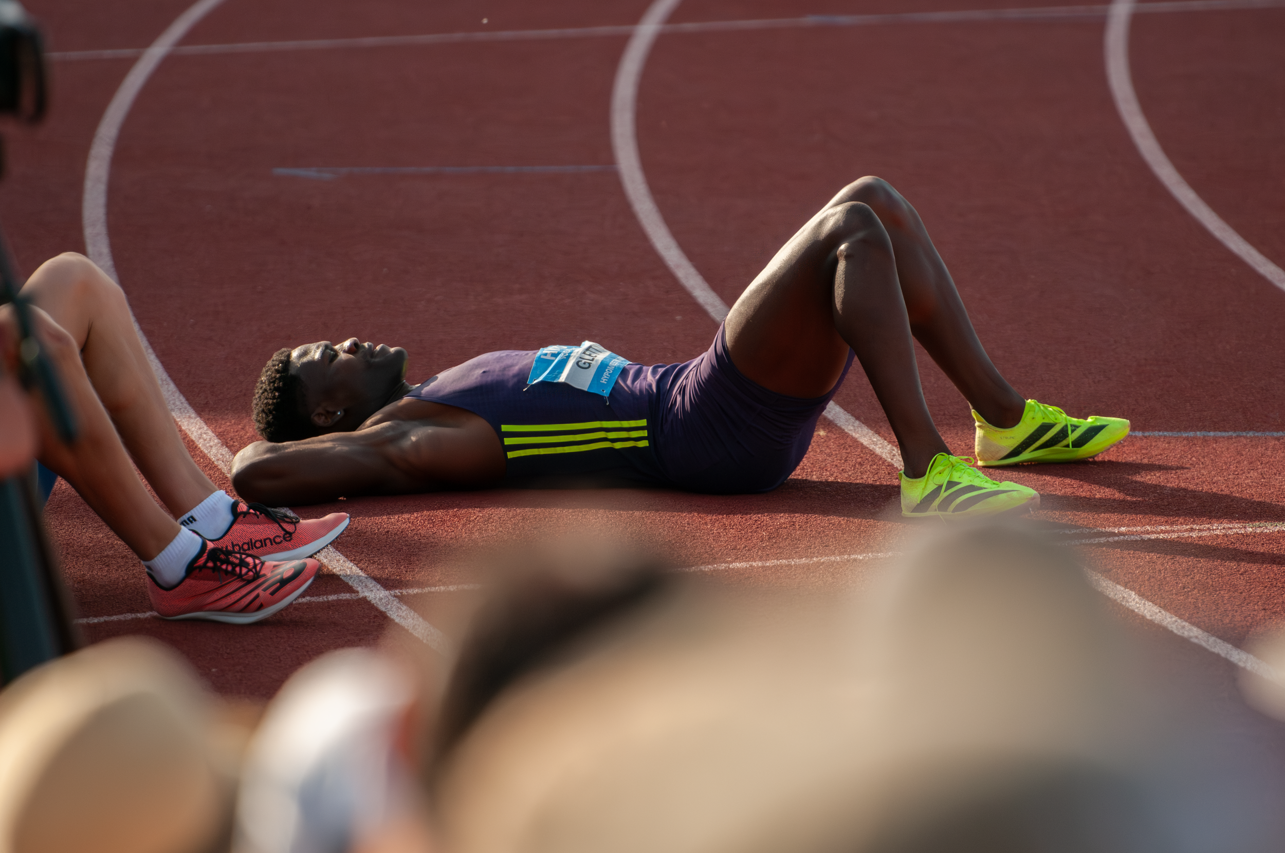 Ein junger männlicher Leichtathlet liegt erschöpft auf einer Laufbahn nach einem Wettkampf, umgeben von anderen Athleten und Zuschauern.