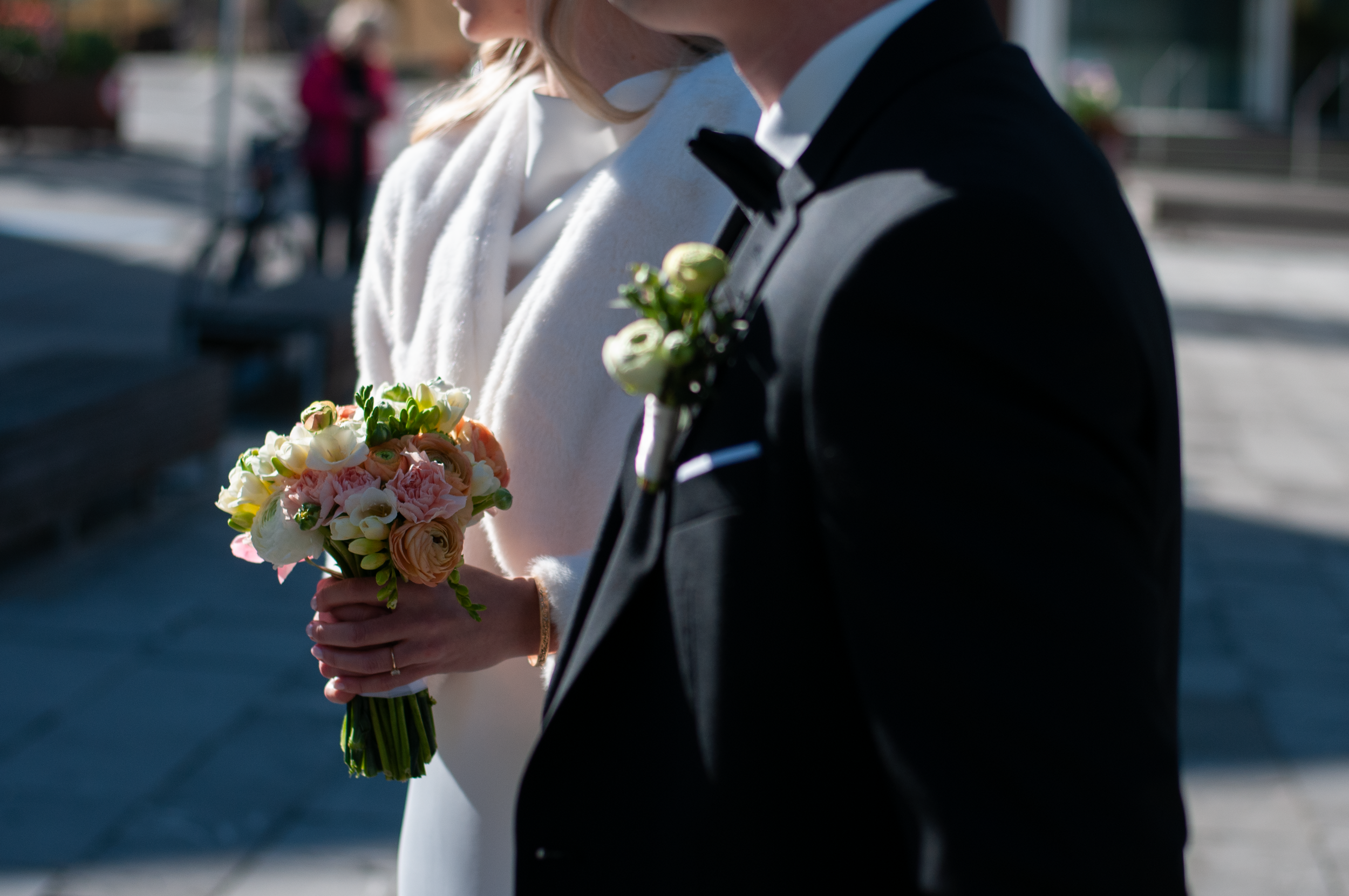 Nahaufnahme eines Bräutigams, der einen Brautstrauß hält bei einer Hochzeit im Freien, im Hintergrund sind unscharf Personen und Gebäude sichtbar.