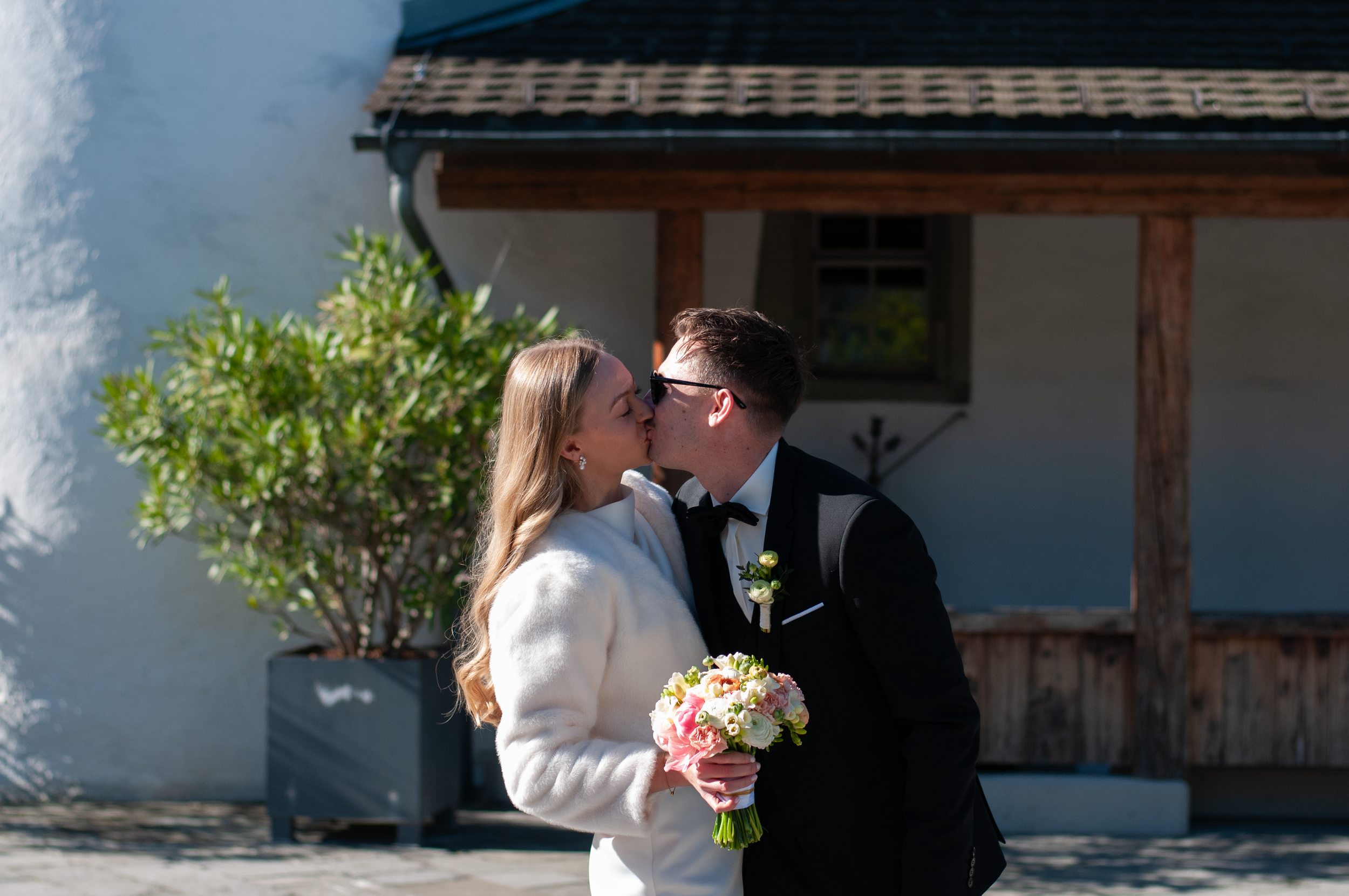 Ein paar küssen sich bei einer Hochzeit im Freien, die Frau trägt ein weißes Kleid und hält einen Blumenstrauß, der Mann trägt einen schwarzen Anzug und eine Schleife.