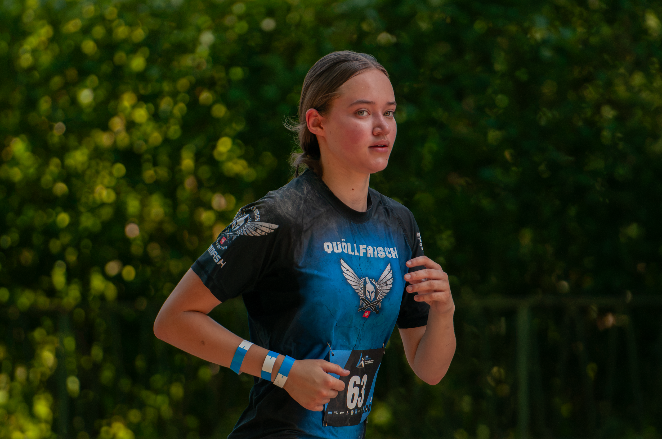 Junge Läuferin bei einem Wettlauf, trägt eine schwarze und blaue Laufshirt mit einem Adler- und Flügel-Logo, eine Nummer 63 auf der Vorderseite, beide Arme mit blauen Bändern, läuft auf einer Strecke mit grünen Bäumen im Hintergrund.