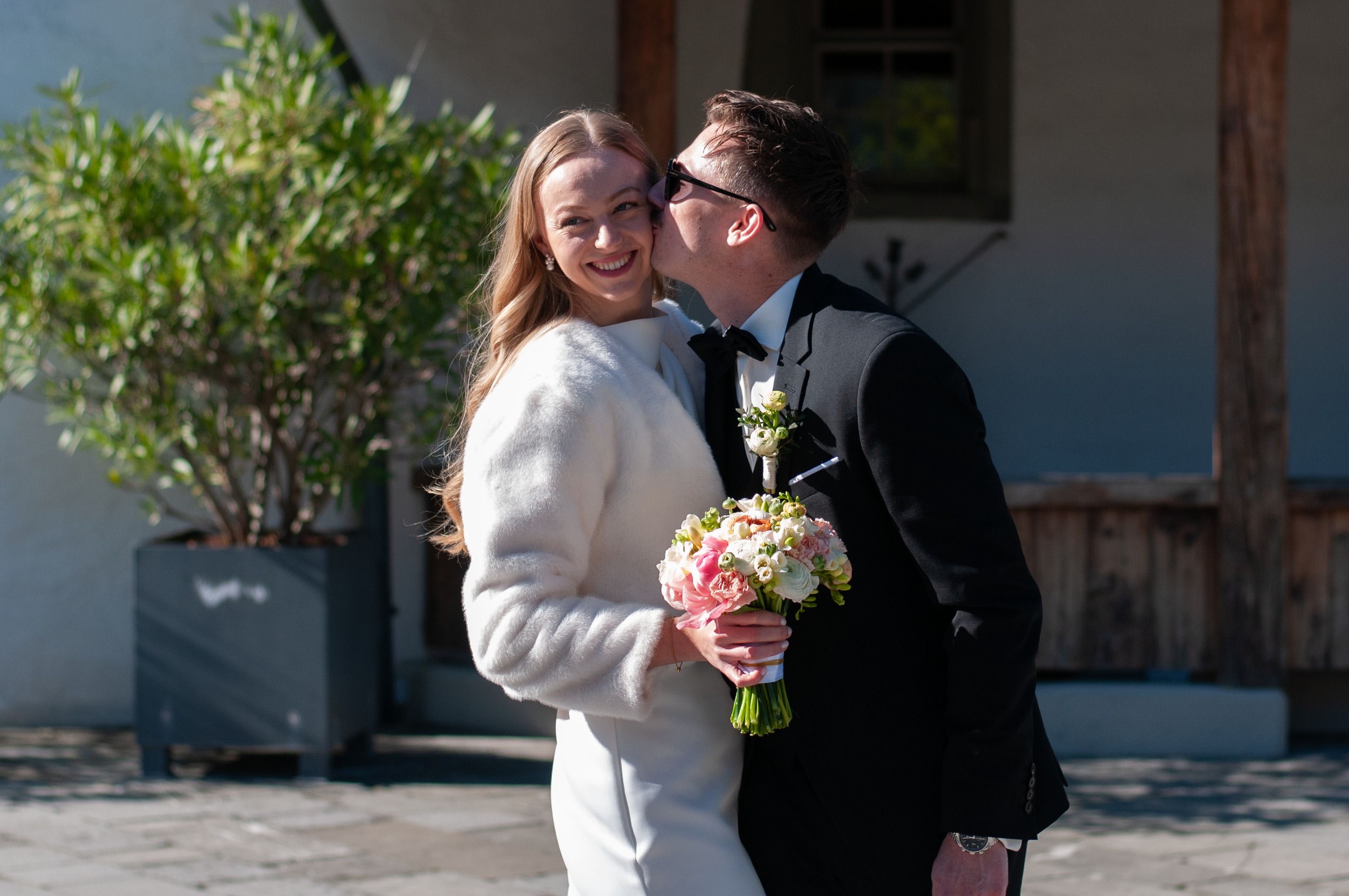 Ein glückliches Brautpaar bei ihrer Hochzeit, sie lächelt und umarmt sich, während er sie küsst. Sie hält einen Blumenstrauß in der Hand. Im Hintergrund sind grüne Pflanzen und ein Gebäude sichtbar.