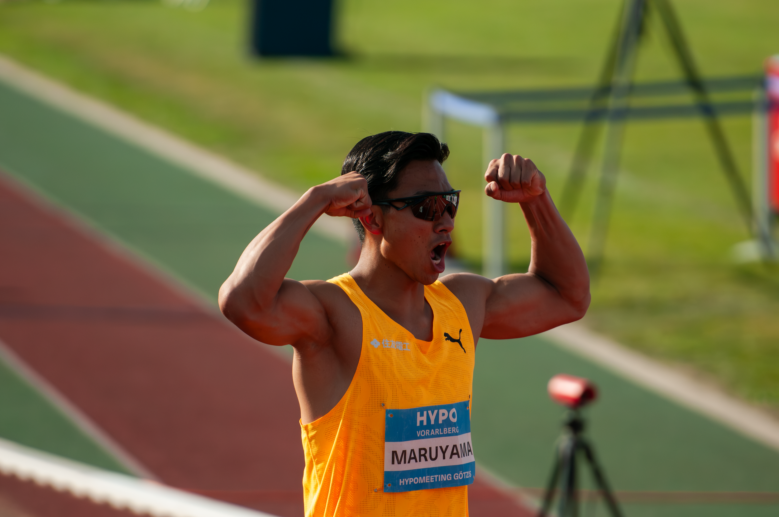 Leistungssportler mit gelbem Trikot beim Wettkampf, zeigt Muskeln, trägt Sonnenbrille, steht auf einer Laufbahn im Freien, Hintergrund ist grün, es handelt sich um ein Leichtathletik-Event im Stadion.