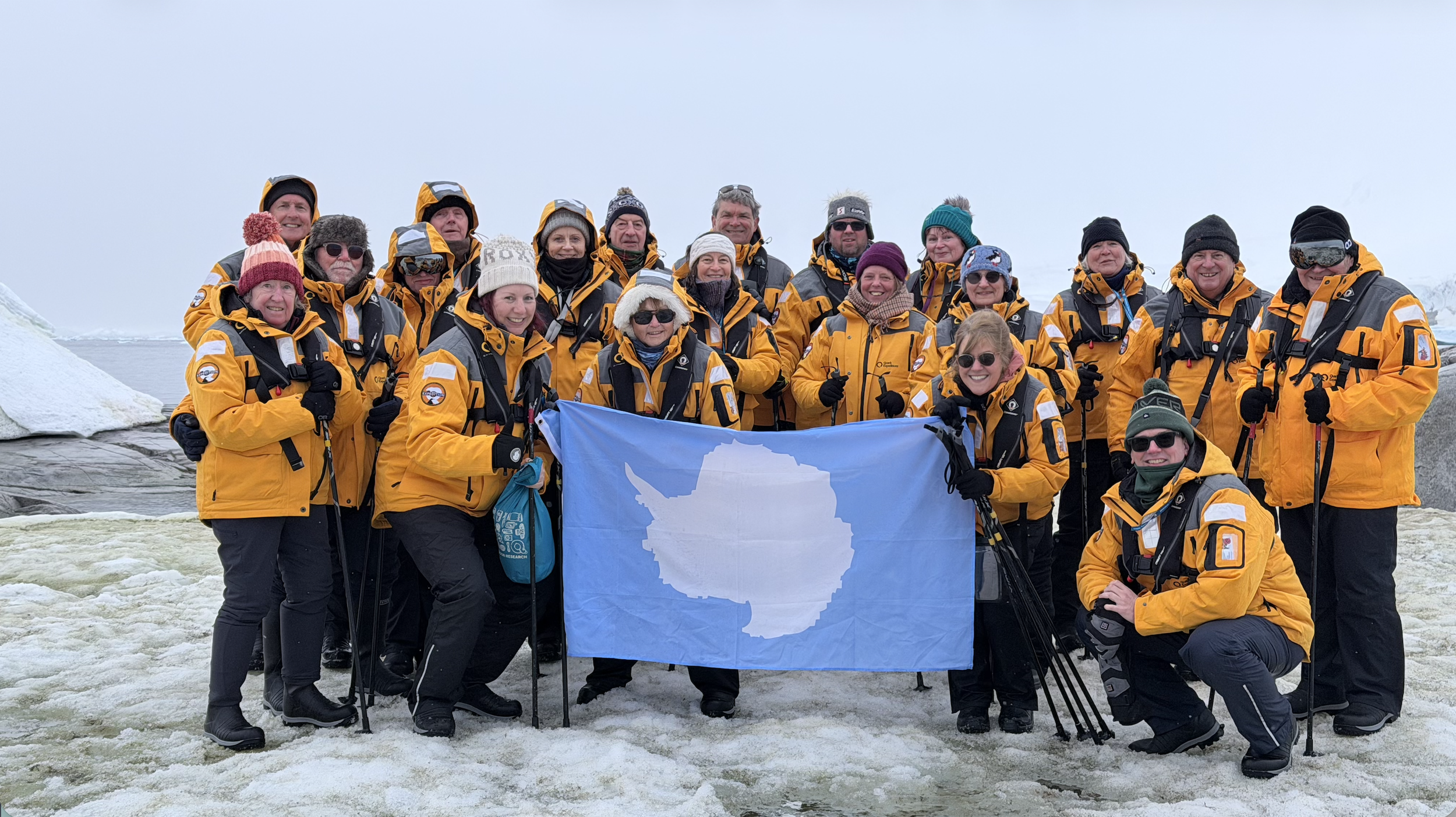 A group of people dressed in yellow and gray winter gear, holding a flag with the shape of Antarctica, standing on icy terrain with snow and ice around them.