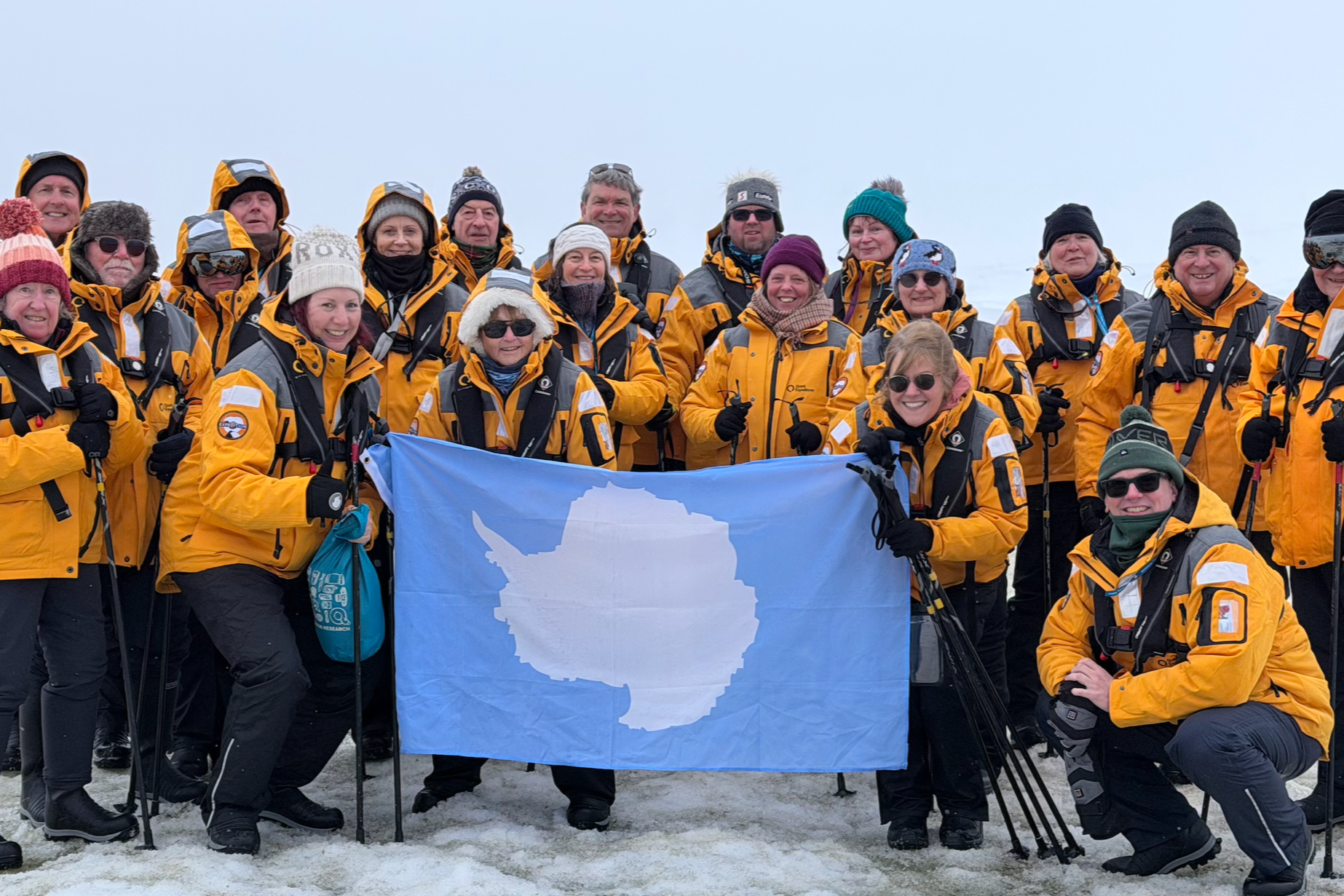 A group of people in yellow and gray outdoor gear on snow, holding a flag with an outline of Antarctica. They are smiling and ready for an expedition or outdoor adventure.