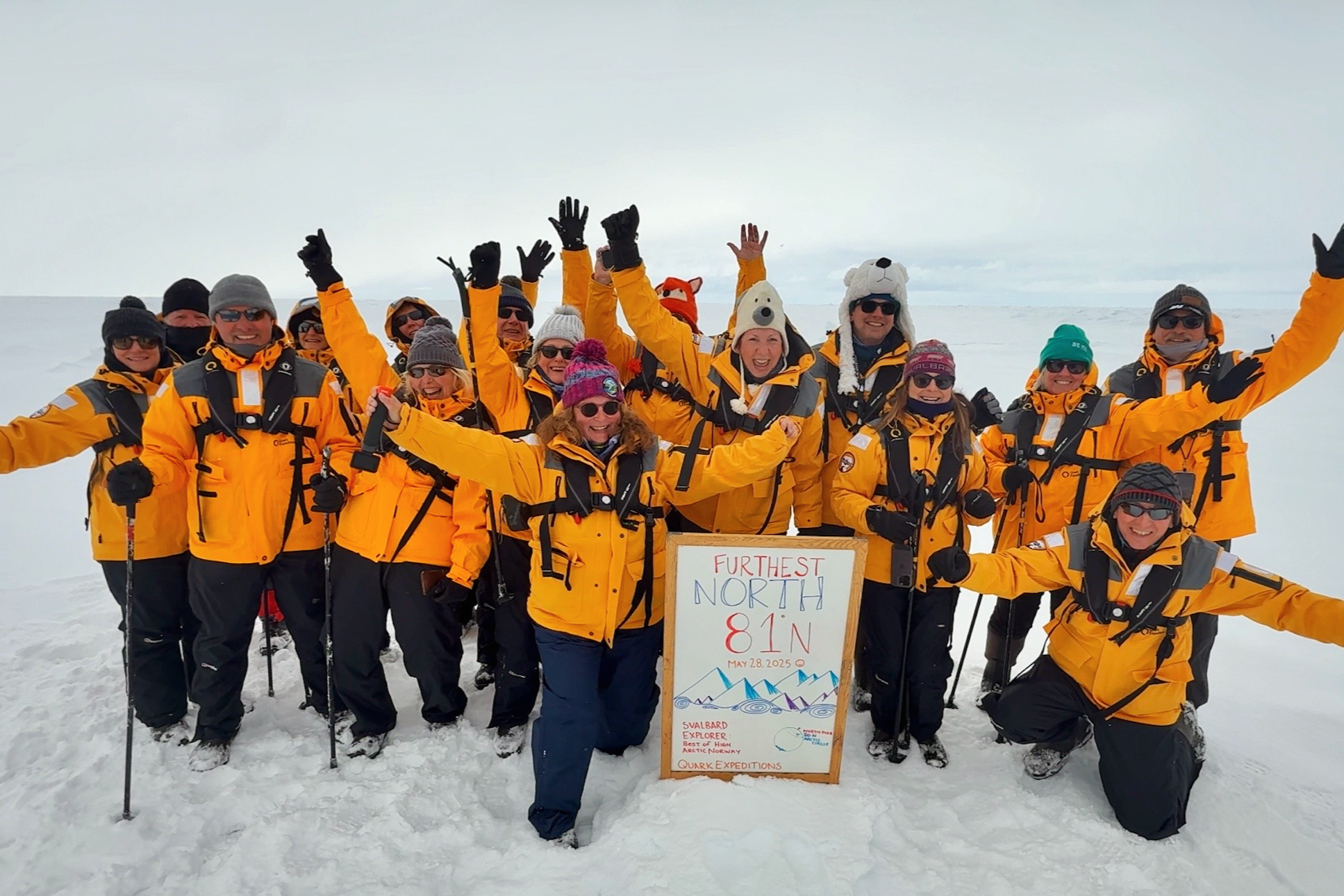 Group of people in yellow jackets and winter gear celebrating on snow-covered landscape, holding a sign that reads "Farthest North 81N" with mountains illustrated on it, and wearing various hats and sunglasses.