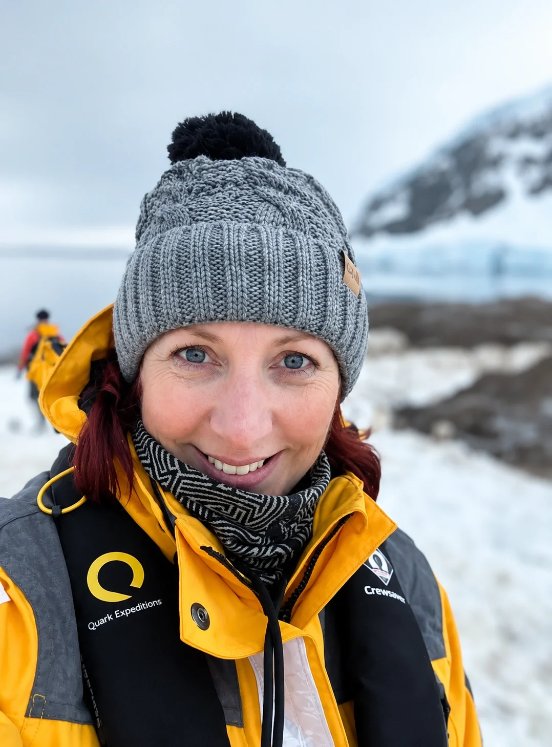 Woman wearing a gray knit beanie and yellow outdoor jacket on a snowy landscape with ice and a mountain in the background.