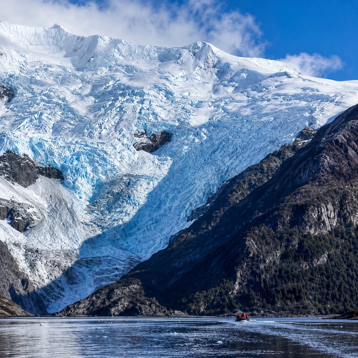 Snow-capped mountain with glacier, dark rocky slopes, and a body of water with a small boat and two people in red jackets.
