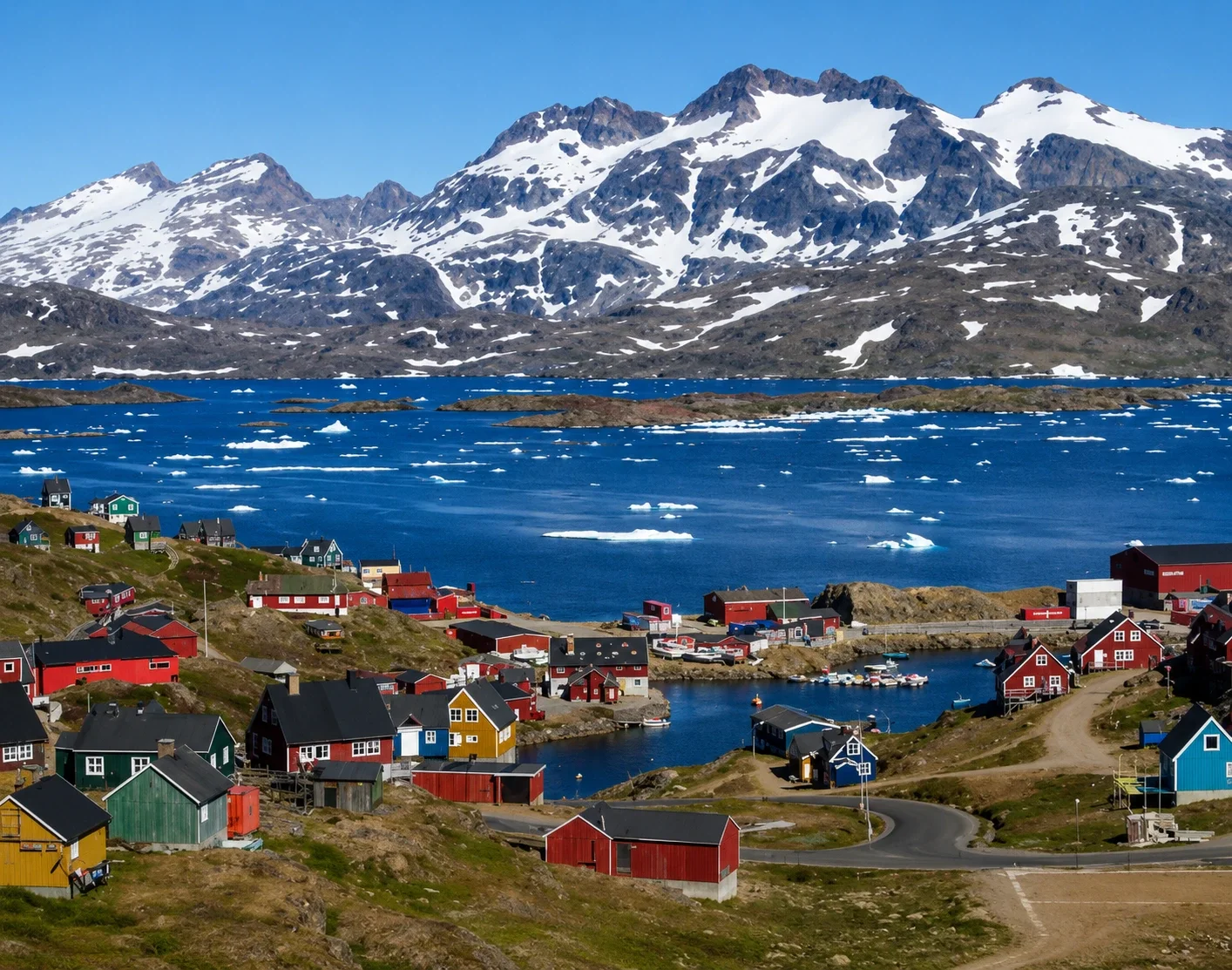 Colorful houses along a coastal village with mountains and snow patches in the background, icy water with floating ice chunks.