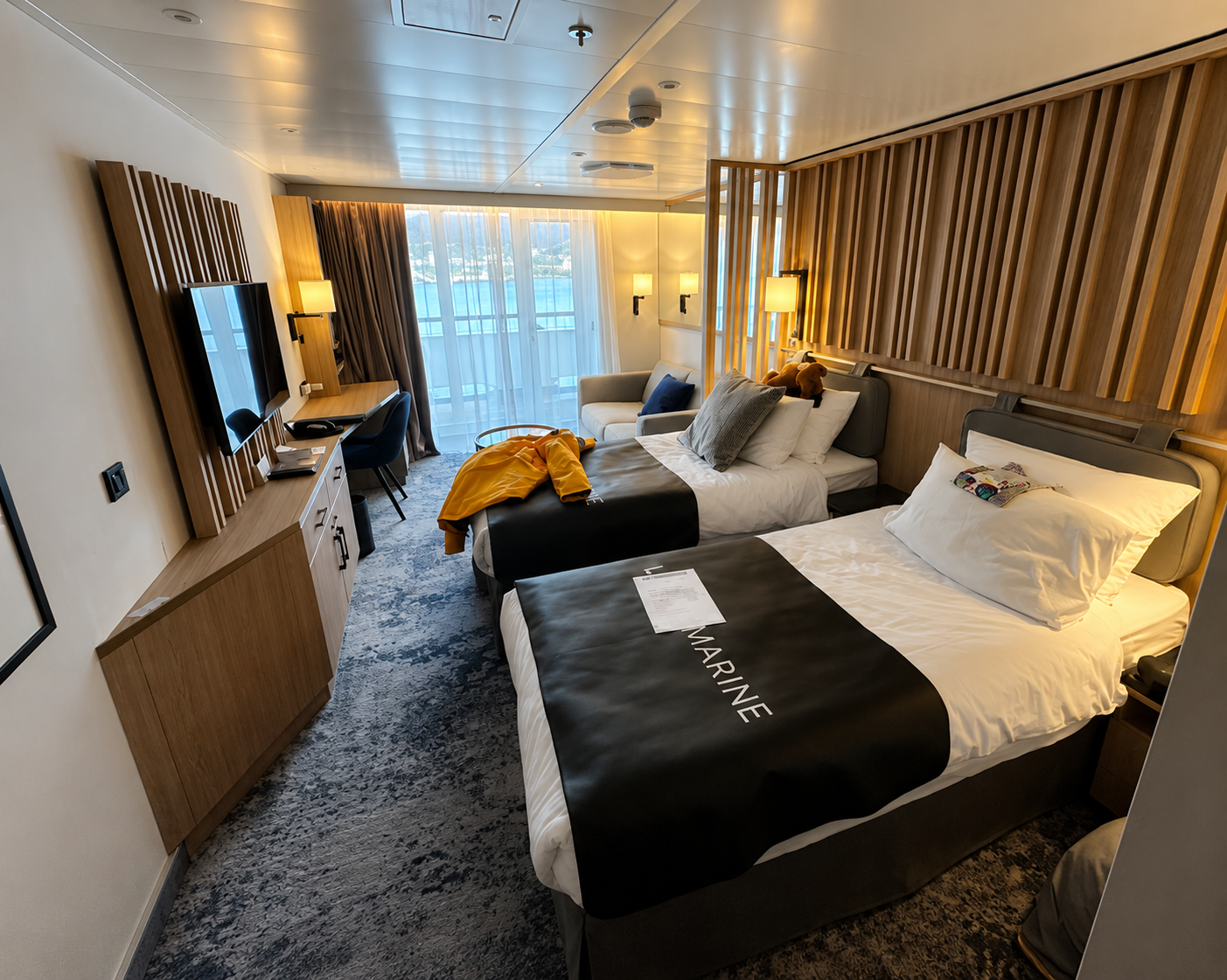A modern hotel room with two beds, one with a gray headboard and the other with a black bed runner labeled 'MARINE'. The room has a large window with sheer curtains, a small sitting area with two chairs and a sofa. There is a wooden partition behind the beds, a TV mounted on the wall, and a desk with a chair near the window.