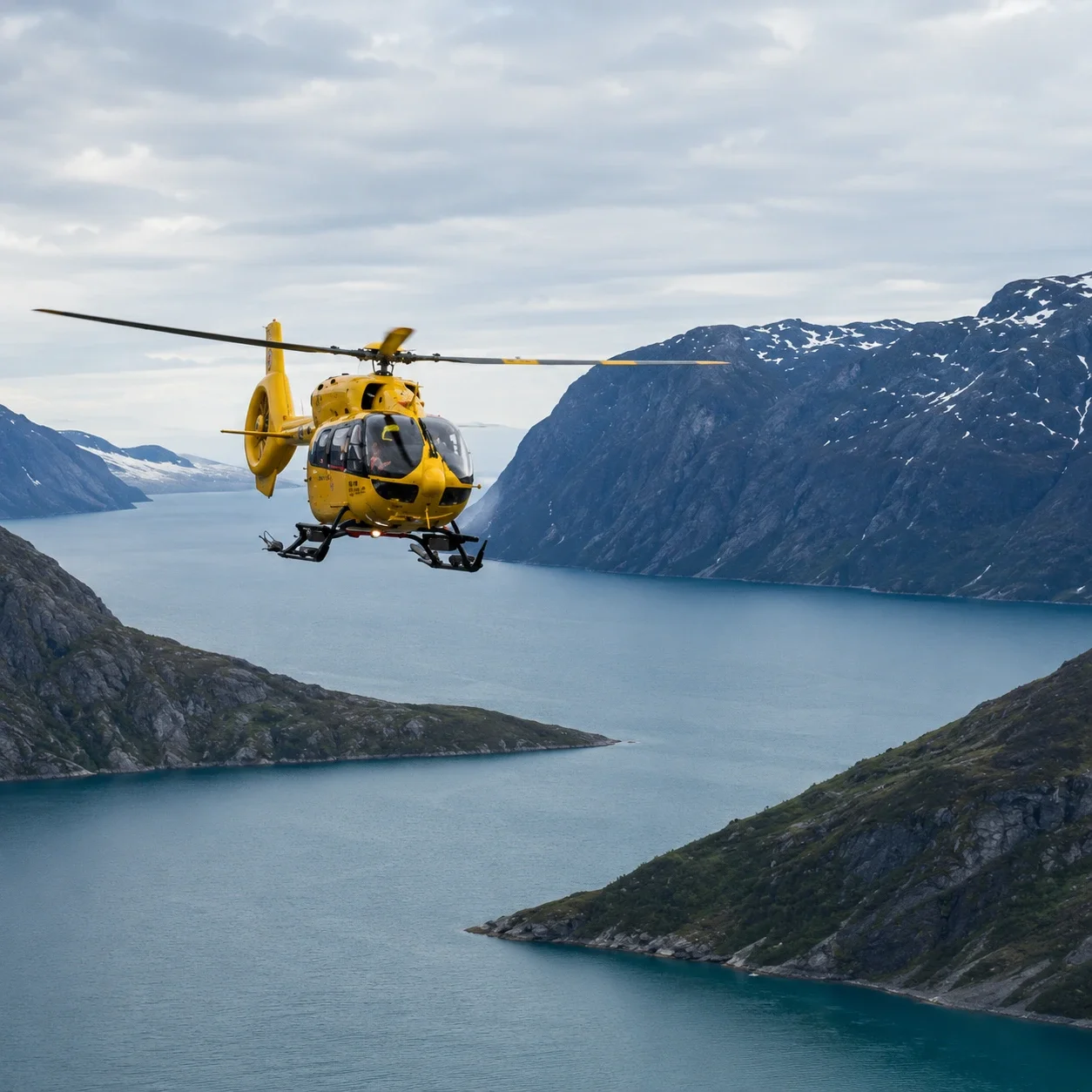 Yellow helicopter flying over a fjord with steep, snowy mountains in the background.