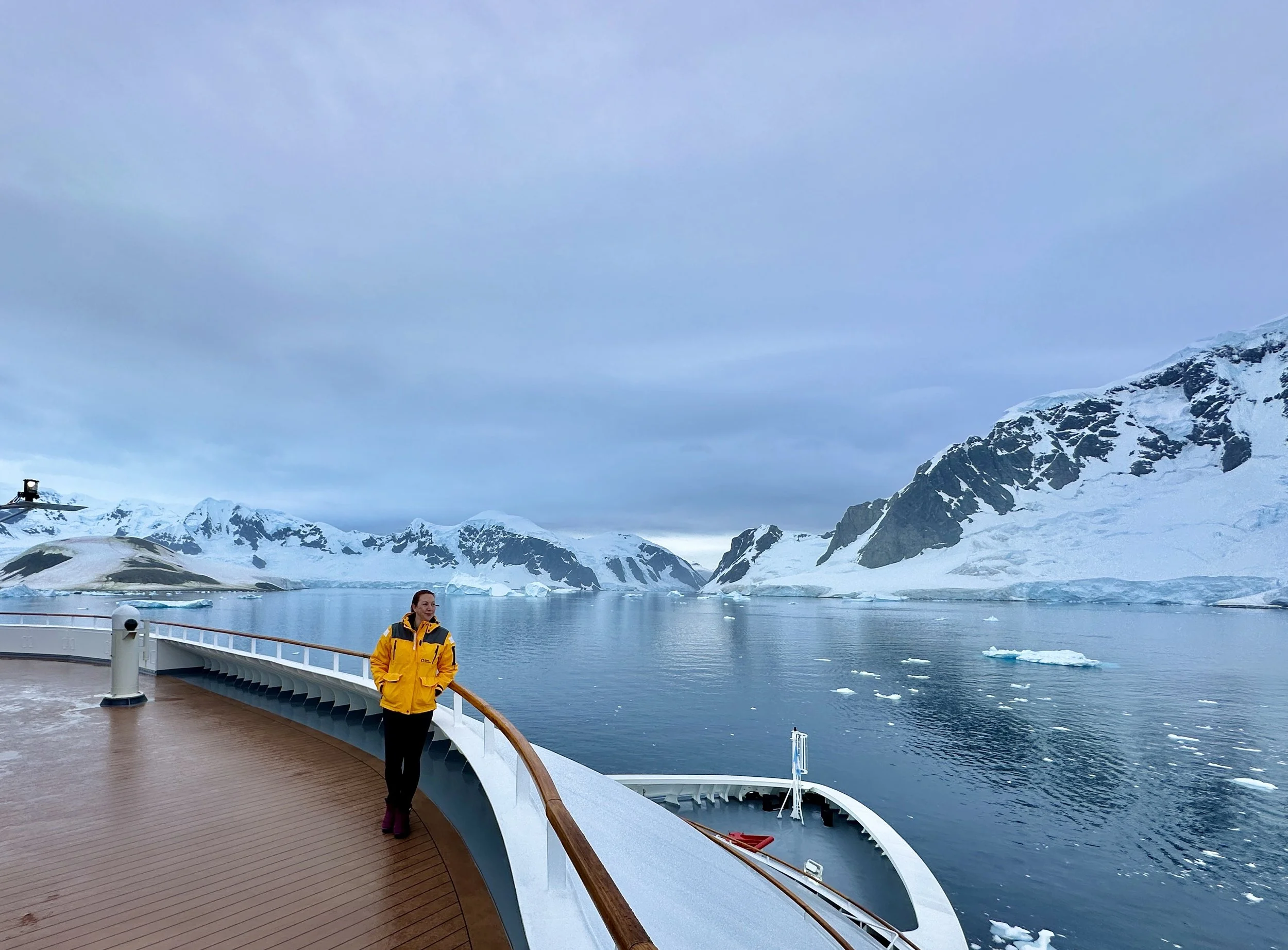 A woman in a yellow jacket standing on the deck of a ship, overlooking icy mountains and icy water with floating icebergs in a cold, cloudy environment.